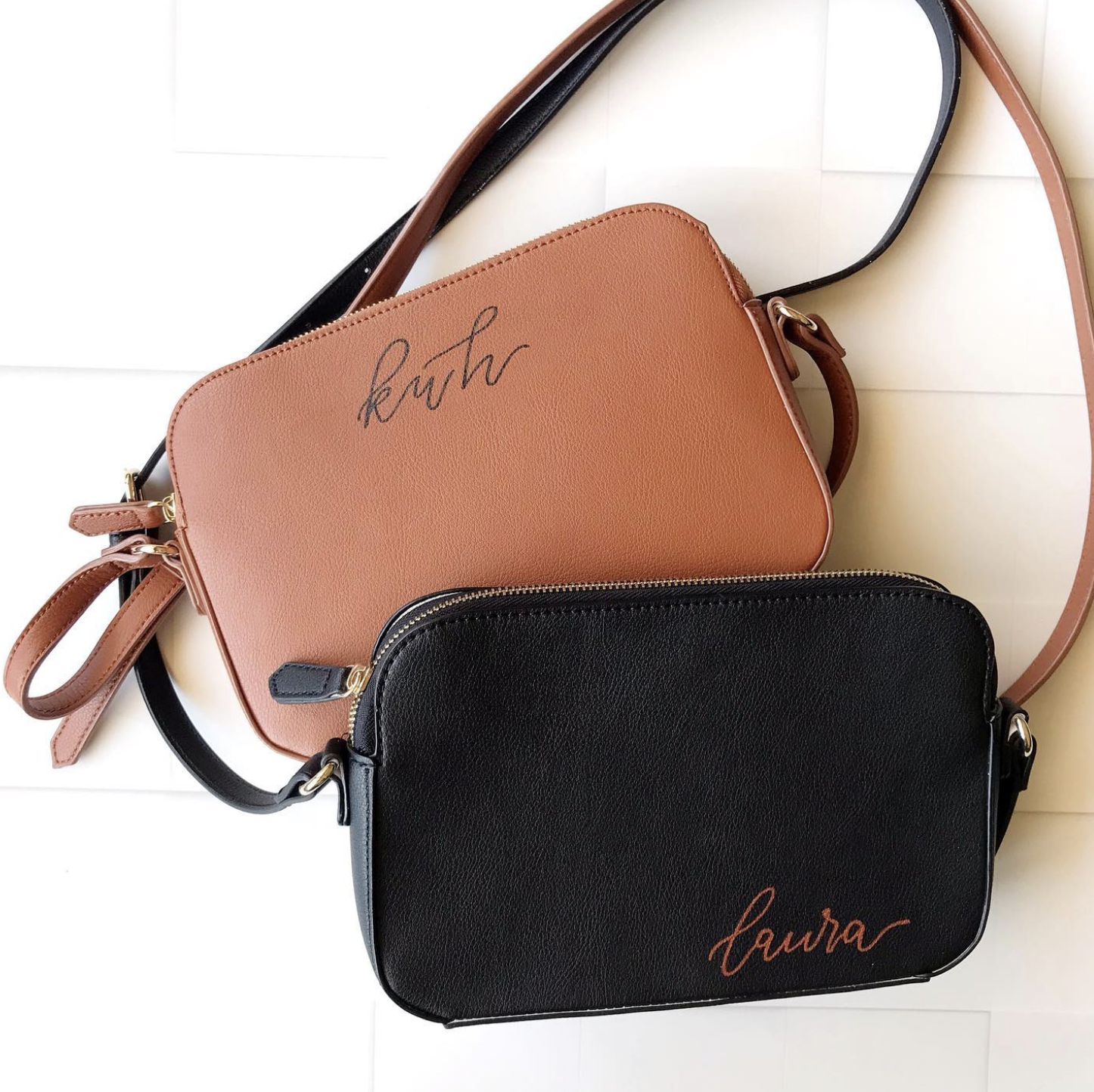 Three leather crossbody bags, one tan with handwritten text, one black with handwritten text, and one black with transparent zipper, arranged on a white wooden surface.