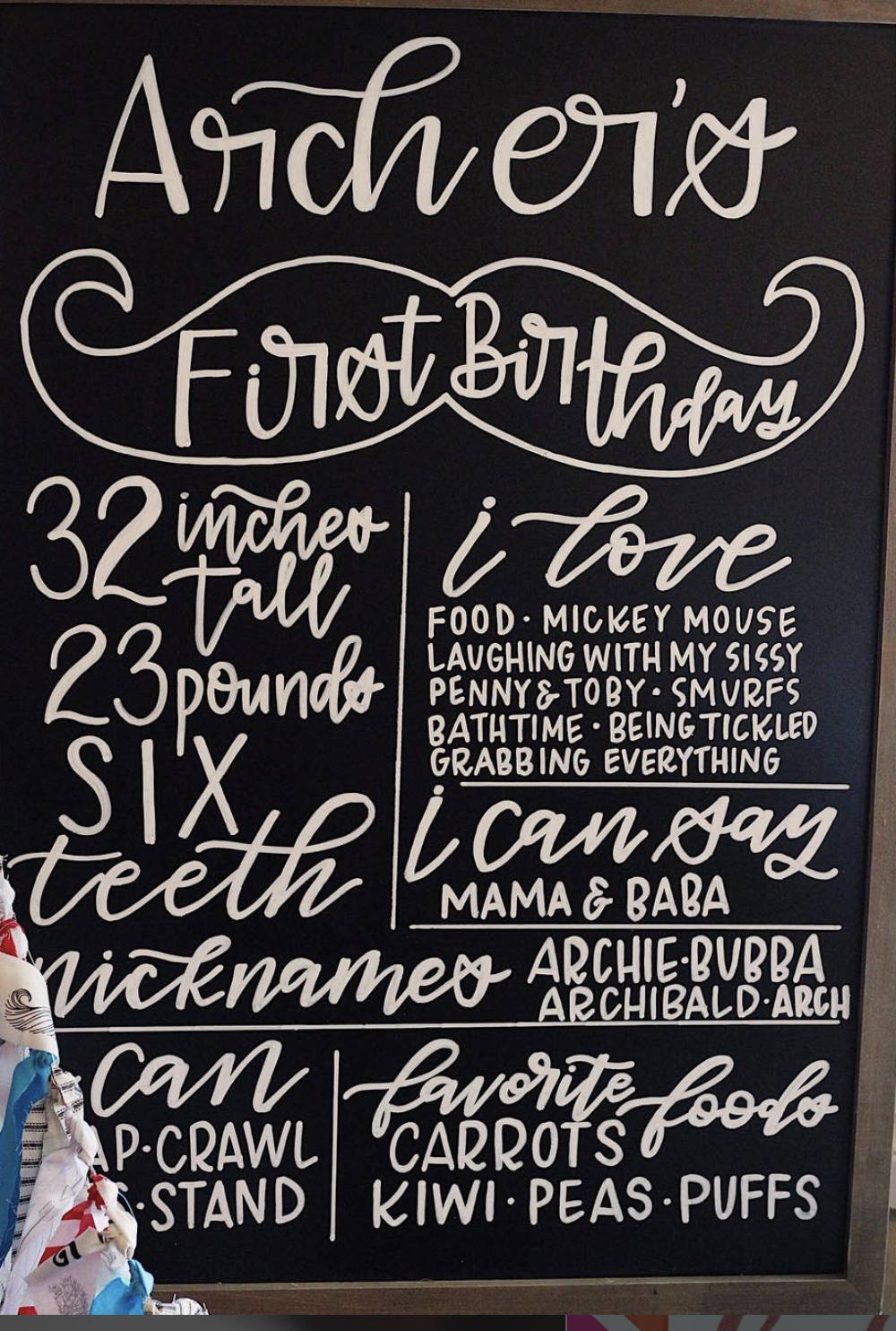 A chalkboard sign celebrating a first birthday with playful and decorative white handwriting. It lists the child's age as 32 inches tall, 23 pounds, and six teeth. The sign includes phrases like 'I love food,' 'I can say mama & baba,' and 'I can craw