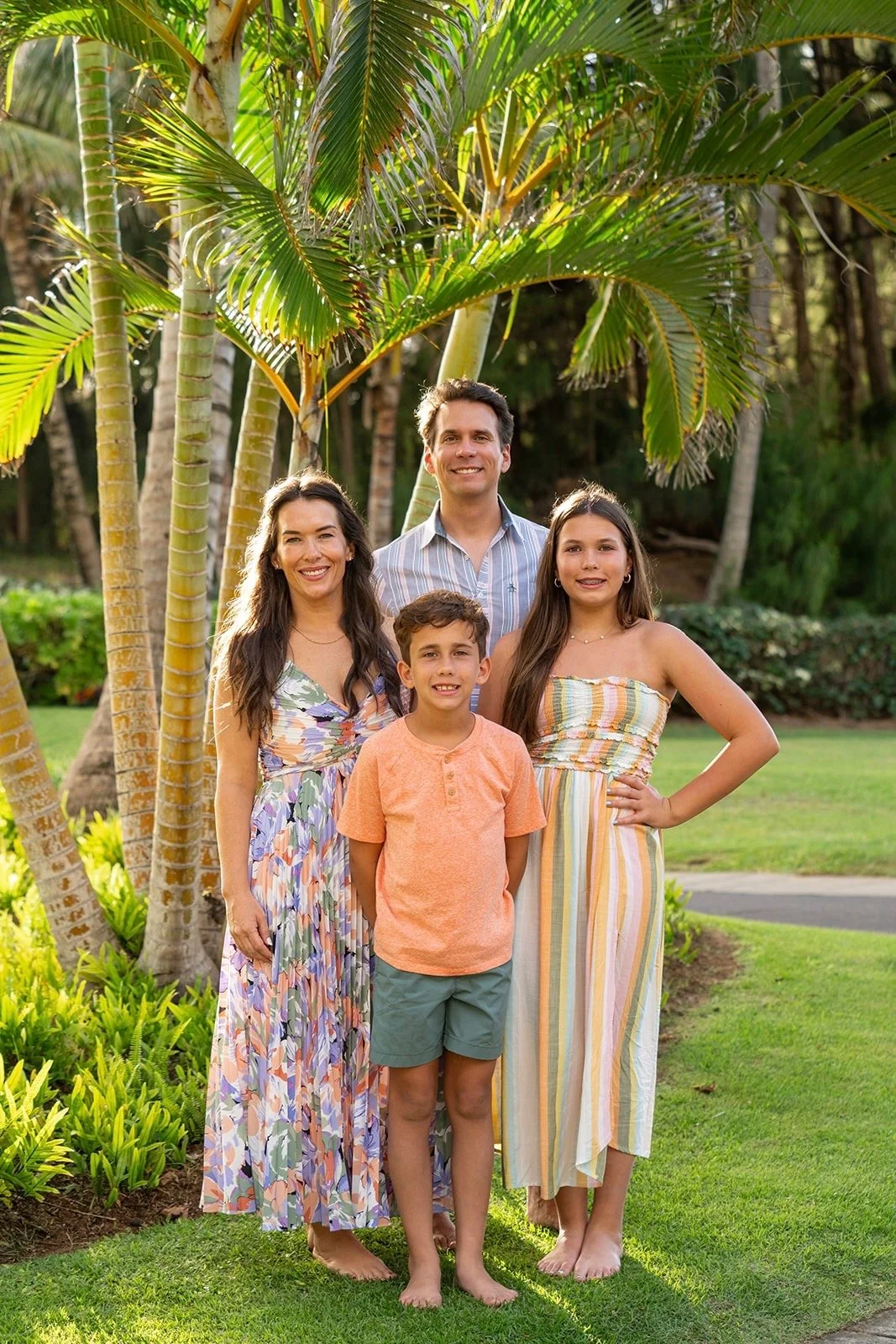 Family of four standing on grass in front of palm trees, smiling, outdoors in warm weather.