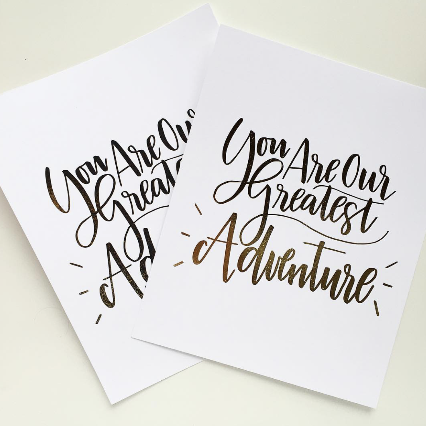 Two sheets of white paper with the phrase "You Are Our Greatest Adventure" written in fancy black and gold cursive lettering.