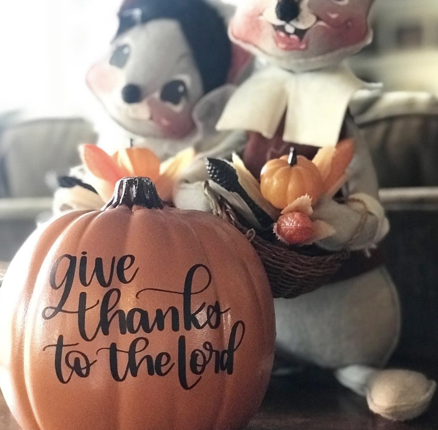 A decorative pumpkin with the words "give thanks to the Lord" written on it, surrounded by small artificial pumpkins and scarecrow figurines.