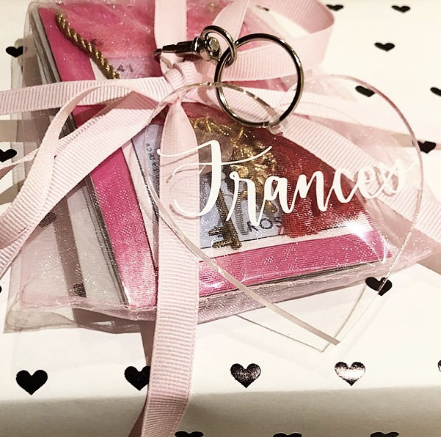 A small gift box wrapped in pink and clear plastic with a pink ribbon and a heart-shaped acrylic ornament with the word 'Mom' on it, tied to the box with a pink ribbon. The box contains a roll of money and some gold jewelry. The background is decorat