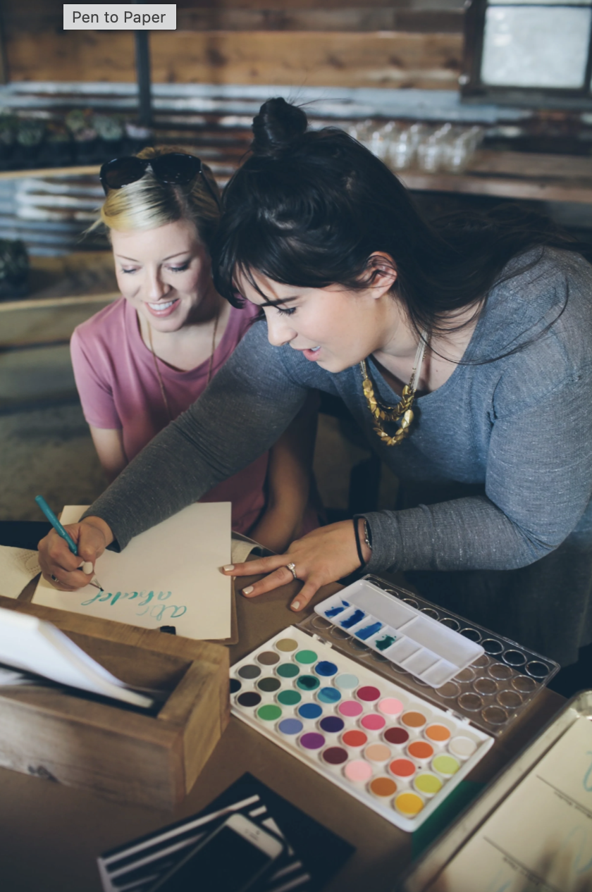 Two women are working together at a table, with one writing calligraphy on a piece of paper. The table has watercolor paints and brushes, and the setting appears to be a cozy, rustic space.
