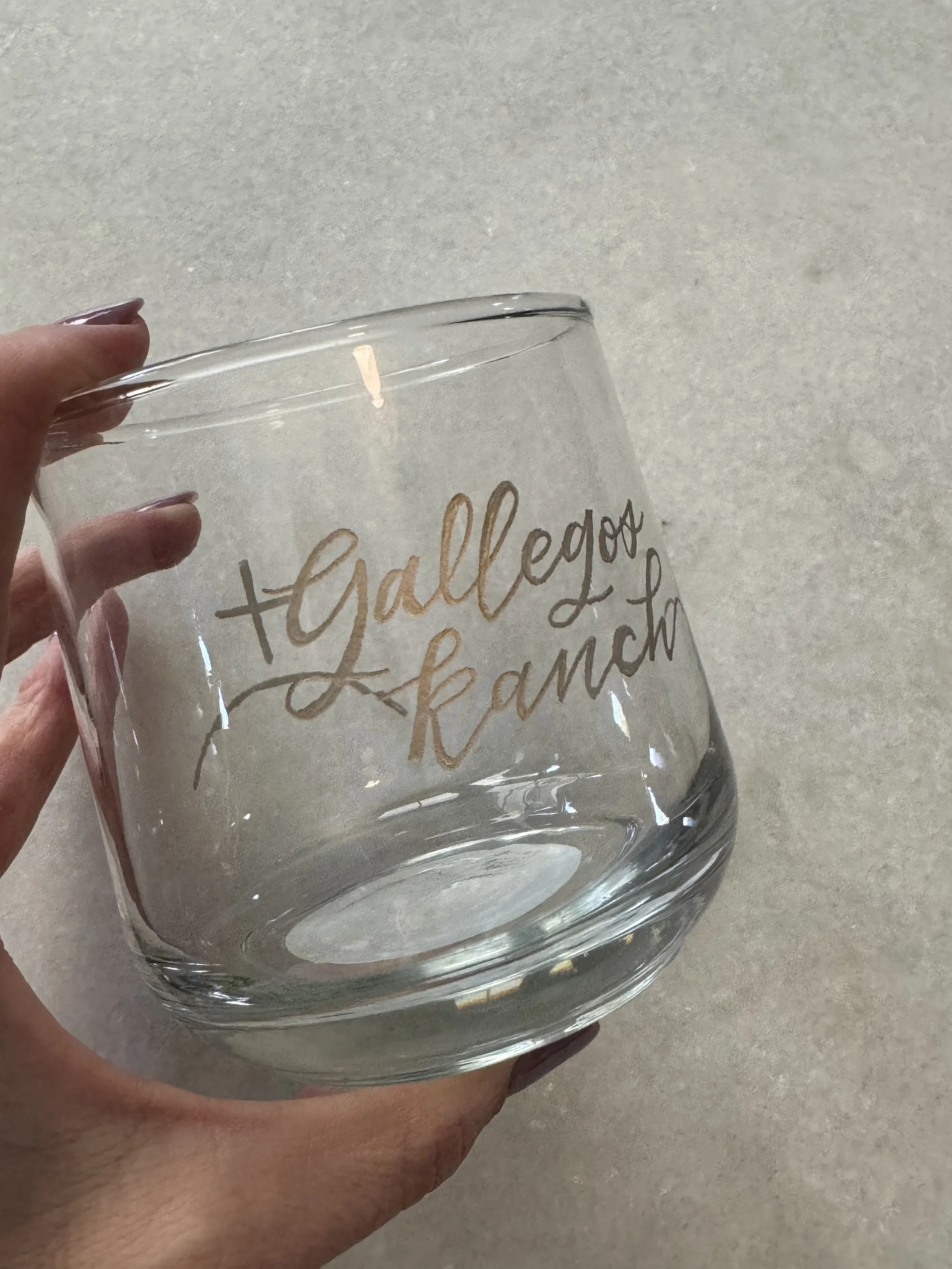 A clear glass cup held in a hand with writing on it that says 'gather' and other words, against a light background.