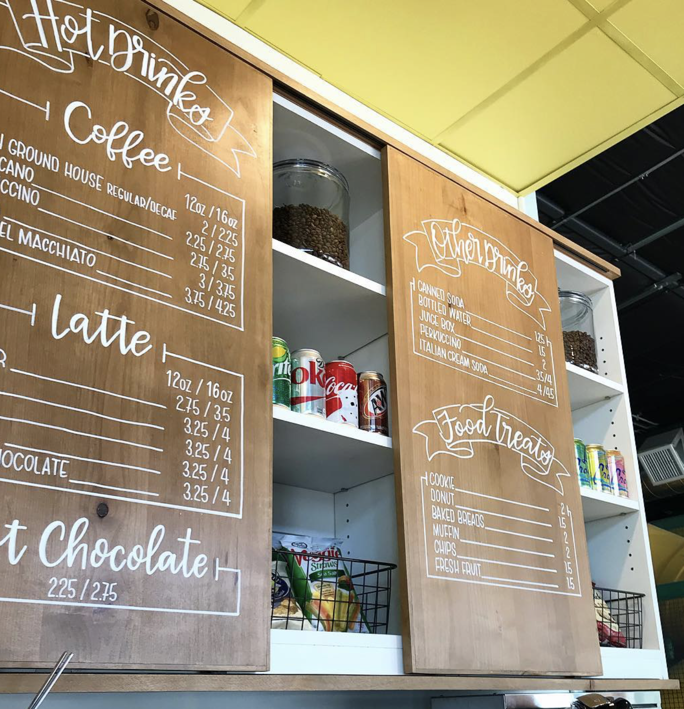 Wooden menu boards with white handwritten text listing coffee, latte, soda, and food items at a café, with shelves of canned drinks, snacks, and coffee beans behind.