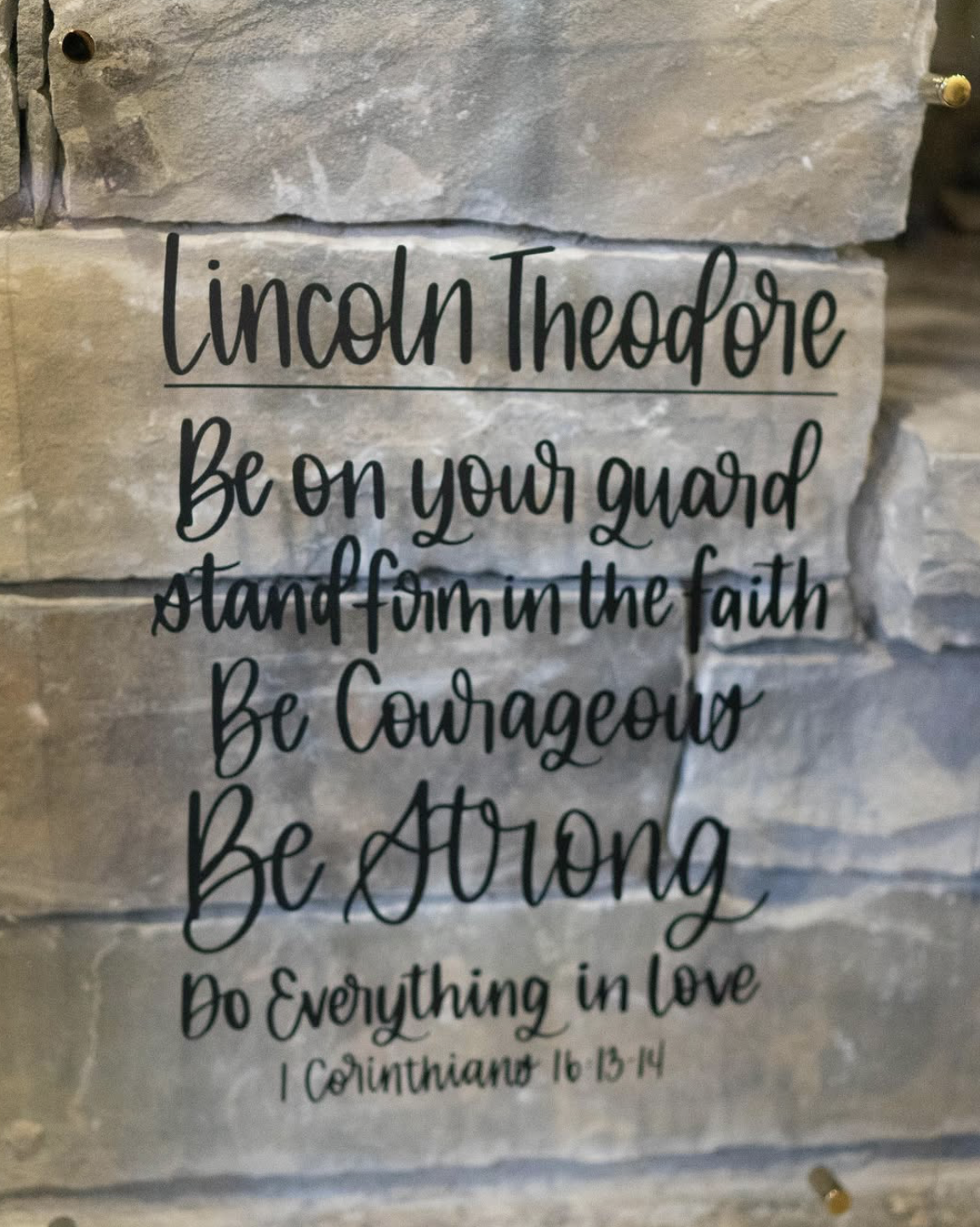 A decorative wall sign with an inspirational religious quote from 1 Corinthians 16:13-14. The sign is mounted on a stone wall and features black cursive text reading, 'Lincoln Theodore. Be on your guard, stand firm in the faith, be courageous, be str