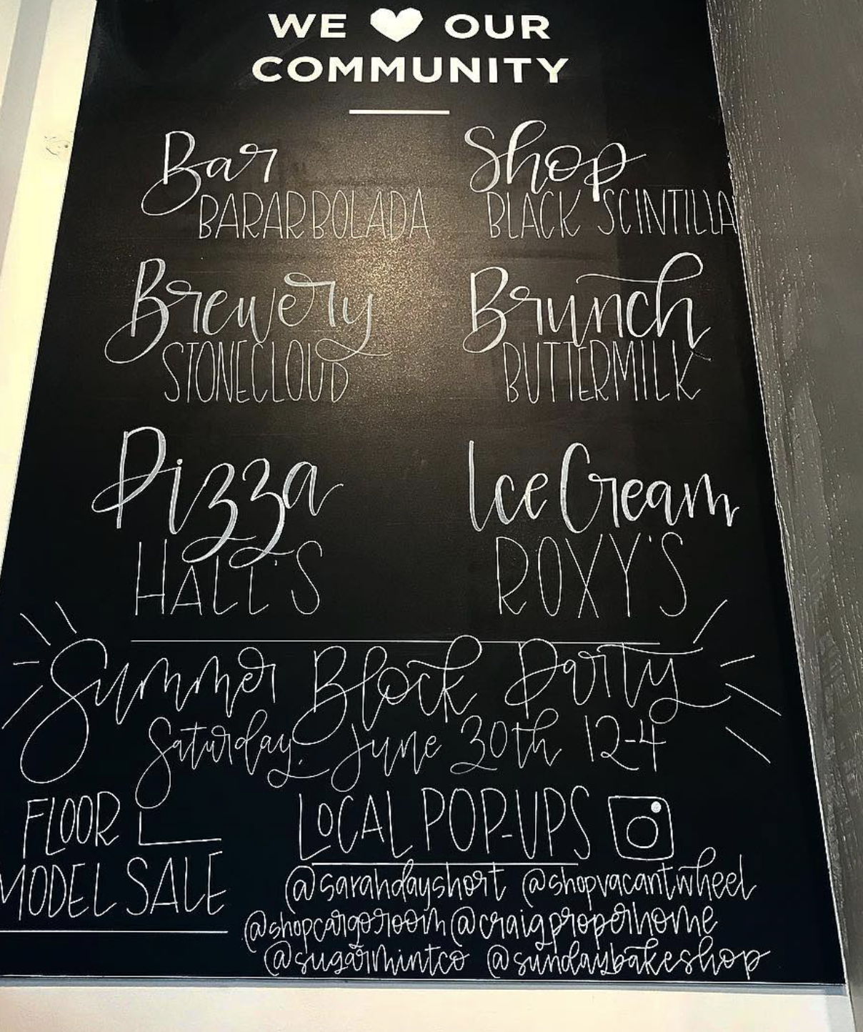 Black chalkboard sign with white handwritten text announcing a community event, including a summer block party on Saturday, June 30th from 12 to 4, with details about food and local pop-ups, and social media handles.