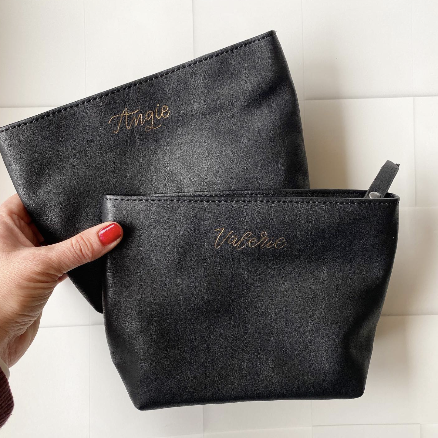 Two black leather handbags with gold embroidery reading 'Vogue' in cursive, one being held by a hand with red nail polish, the other placed on a white surface.