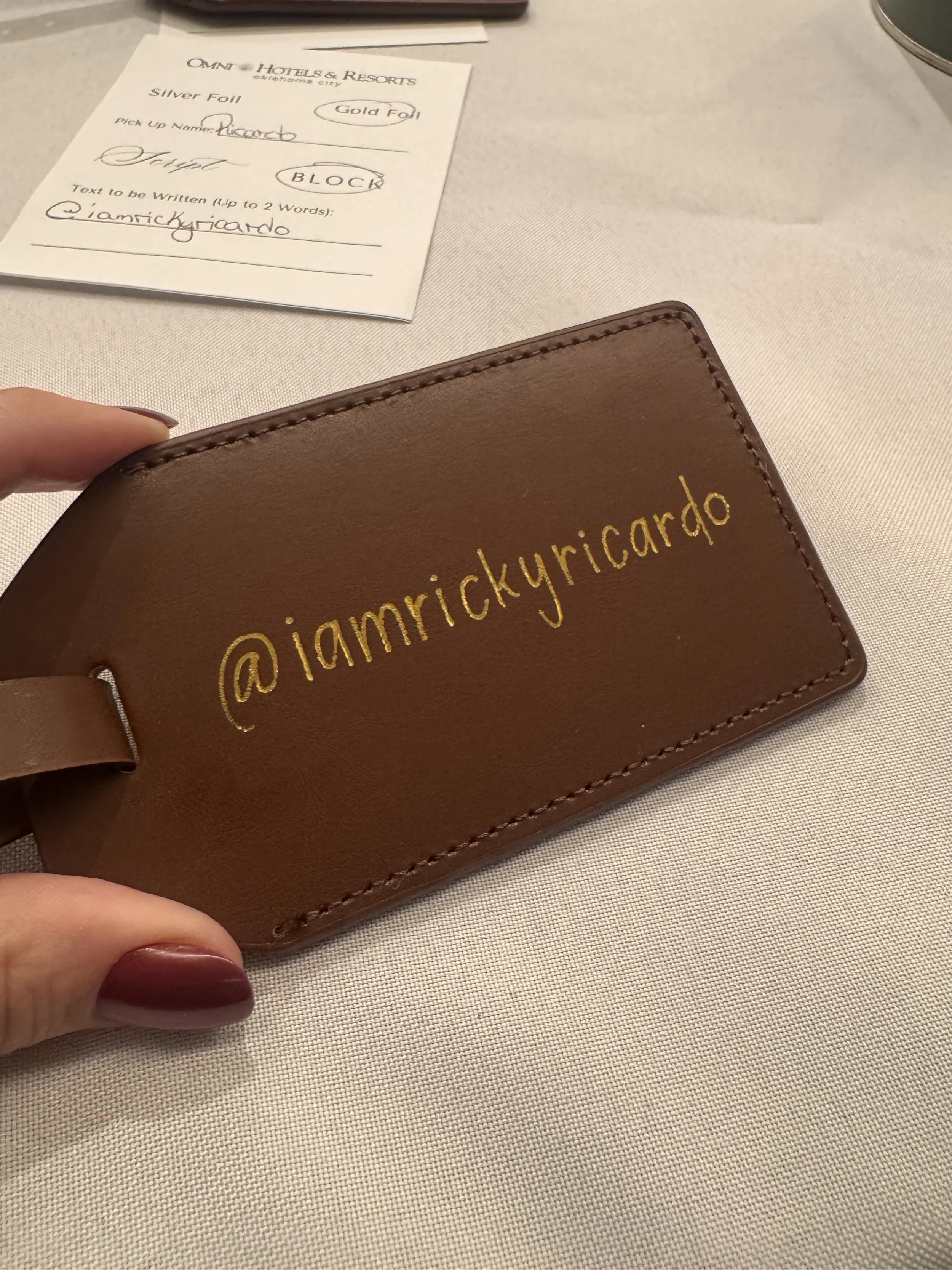 Close-up of a brown leather cardholder with yellow embroidered Instagram handle '@iamrickyricado'. In the background, a hotel check-in slip on a white table with some handwritten information.