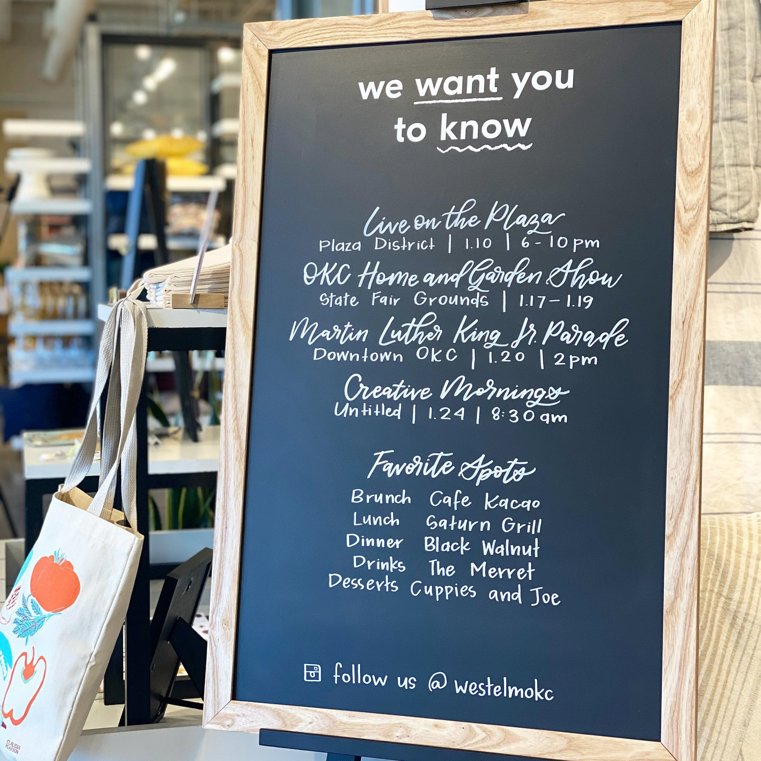 A chalkboard sign with a wooden frame displaying a list of events and activities, including live music, a home and garden show, and various local parades, along with social media follow instructions at the bottom.