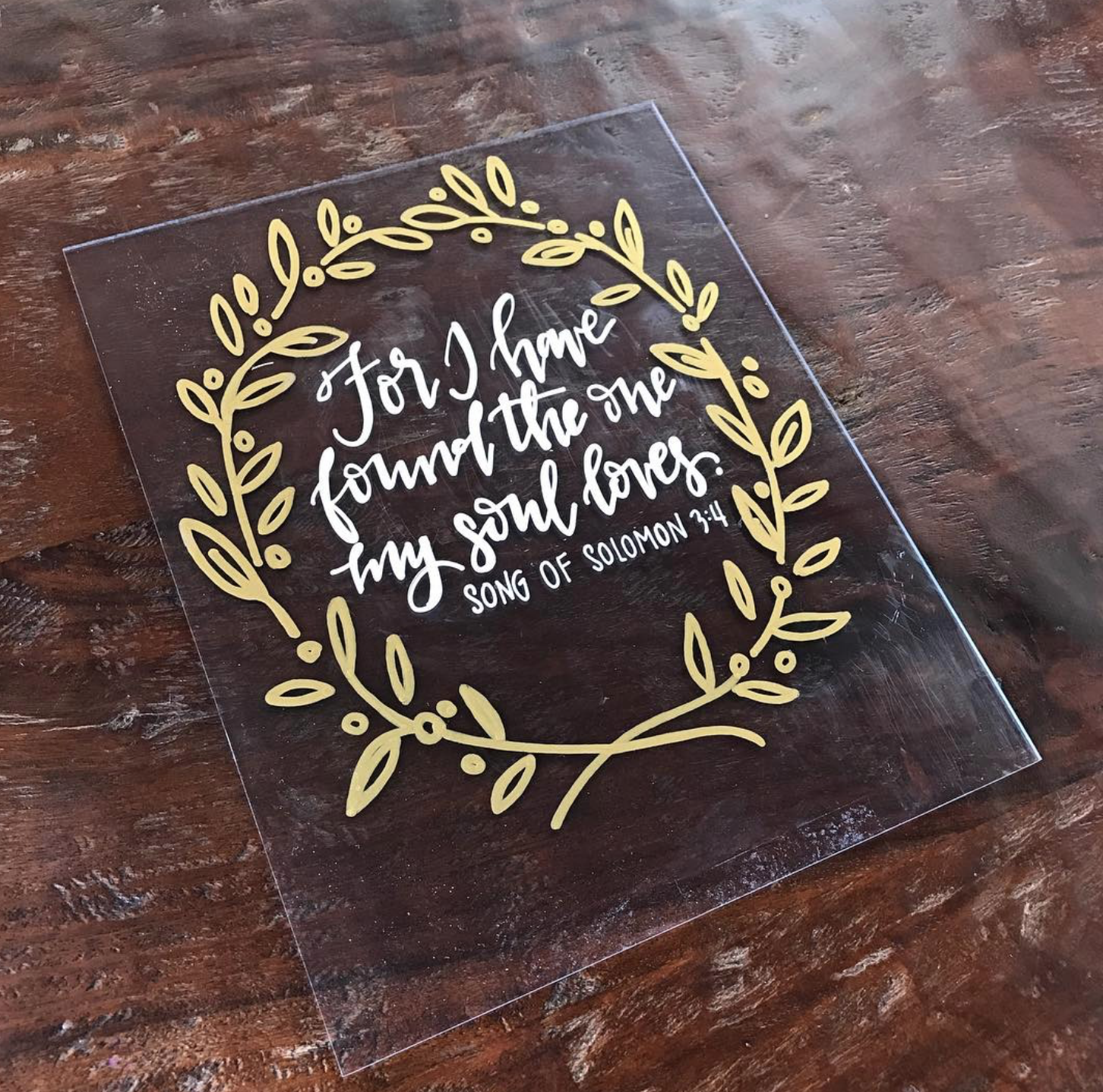 A decorative sign on a wooden surface with a quote from Song of Solomon 2:4, surrounded by a gold leafy wreath design.