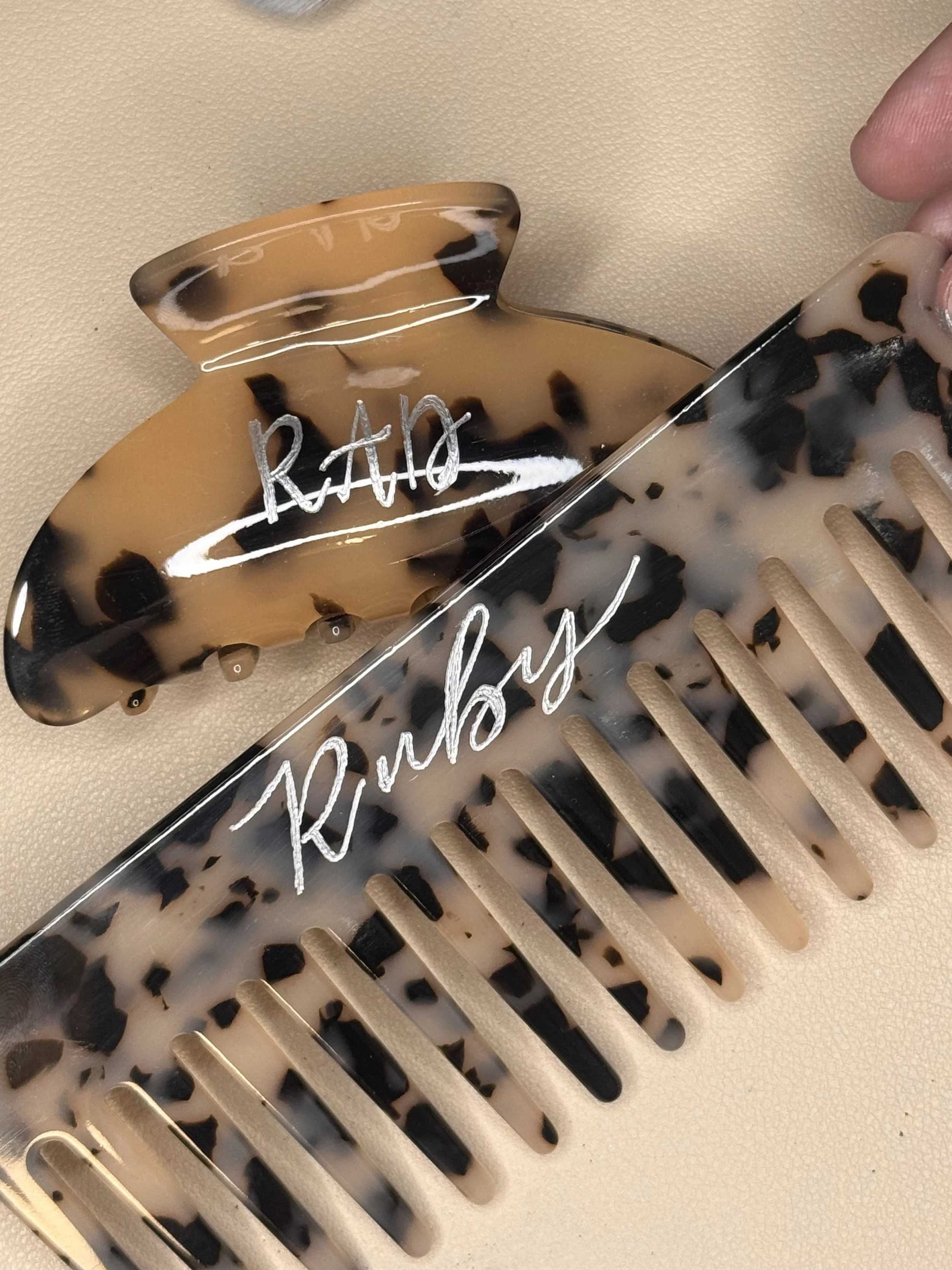 A beige and black tortoiseshell hair clip with white handwritten text on it, partly visible, held by a person's fingers.