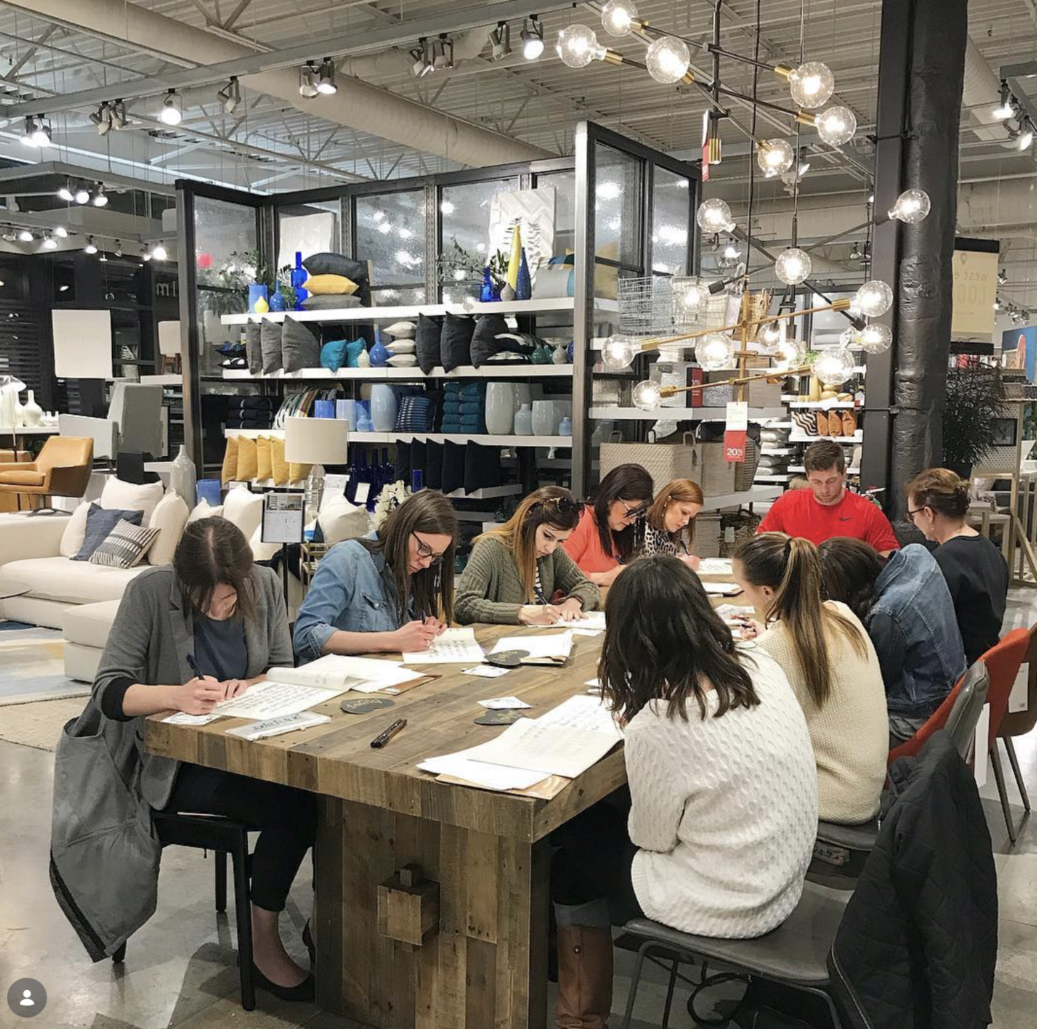 A group of people sitting around a large wooden table, studying or working with papers and notebooks, inside a furniture store with shelves of decorative items and lighting fixtures in the background.