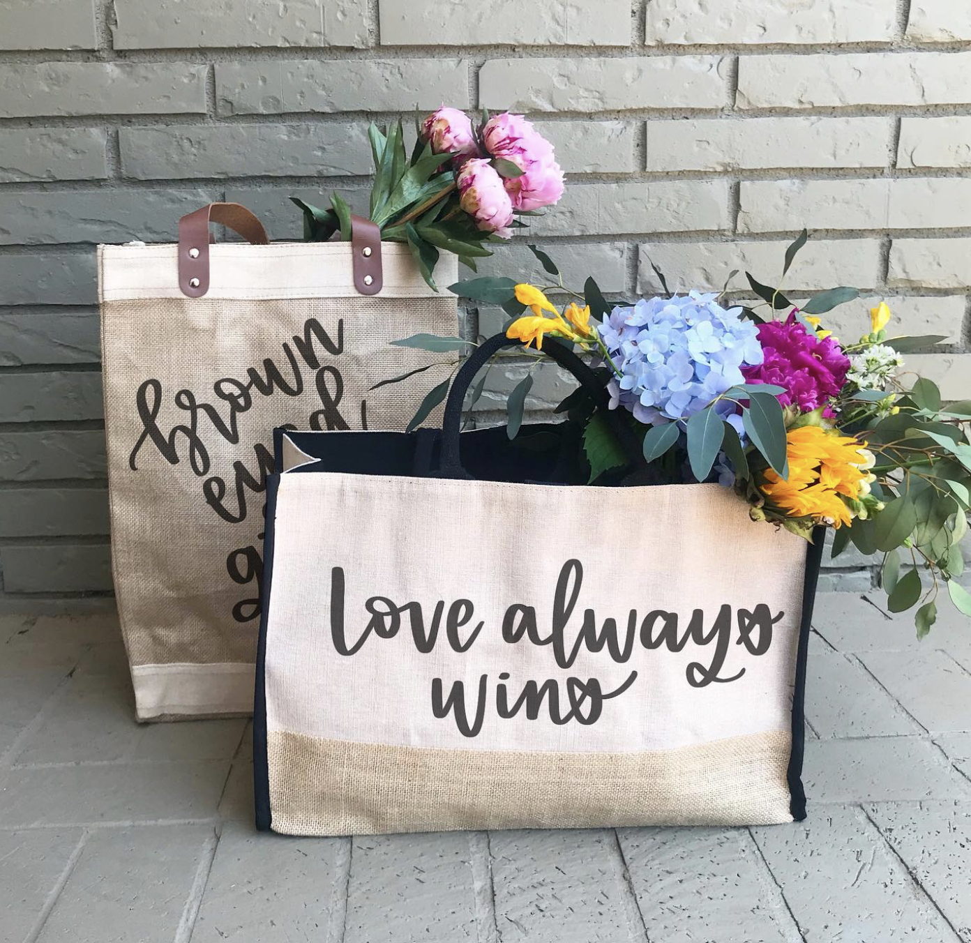 Two tote bags filled with colorful flowers, one with pink peonies and the other with purple, yellow, and white flowers, placed against a gray brick wall. The tote bags have inspiring quotes, with the front bag saying "love always wins" and the back b