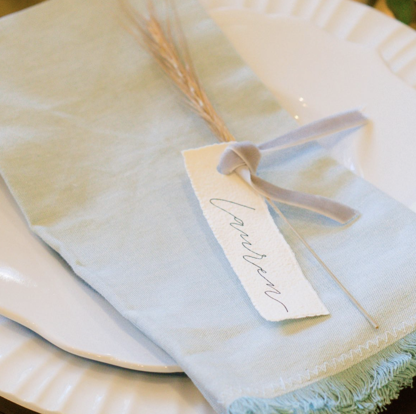 A close-up of a neatly folded white napkin with a small white tag that has the word 'Simple' written on it, secured with a white ribbon, placed on a white plate.
