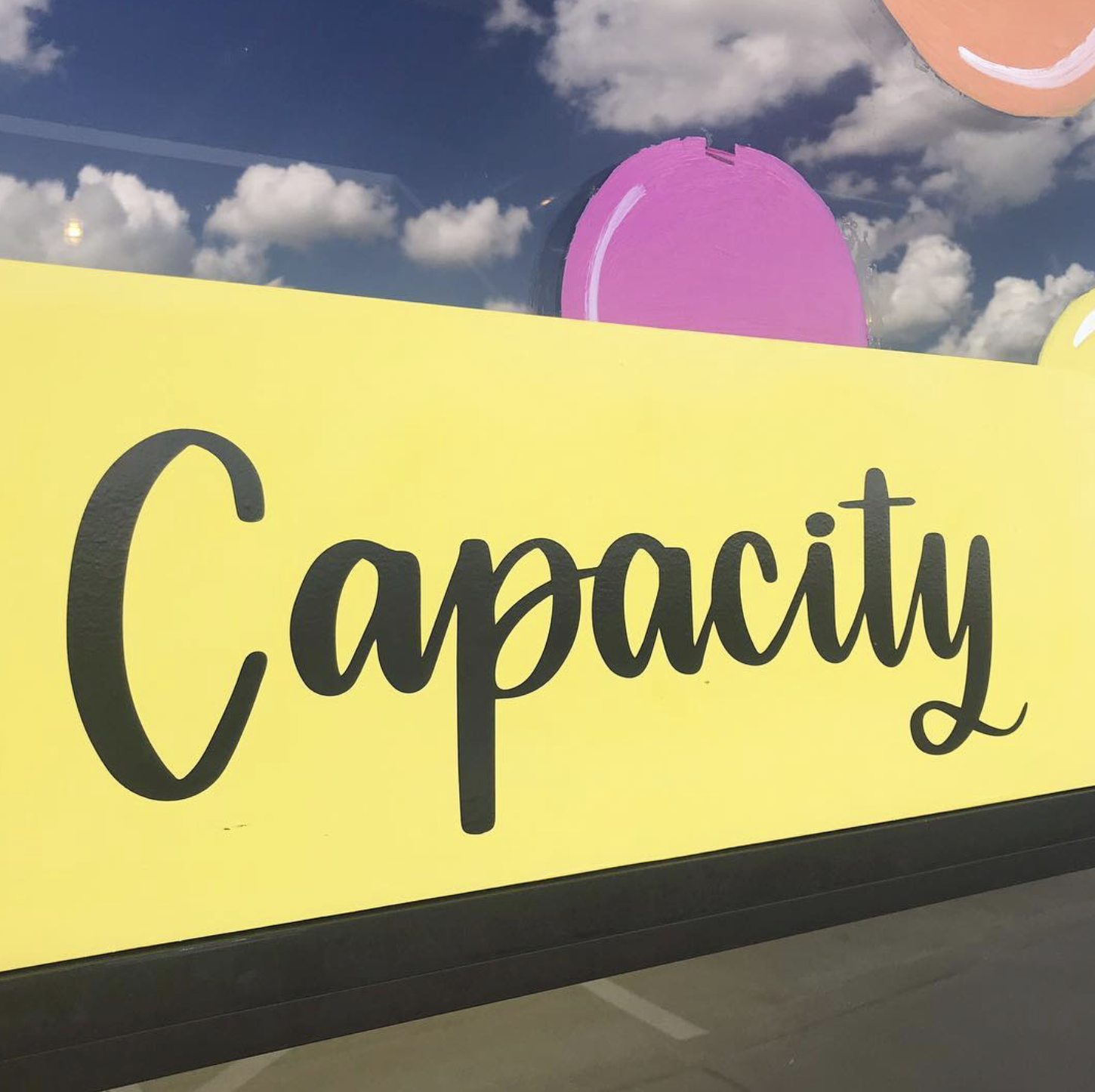 A yellow sign with the word 'Capacity' written in black cursive font, displayed in front of a background showing a blue sky with scattered white clouds and part of a pink circular object.