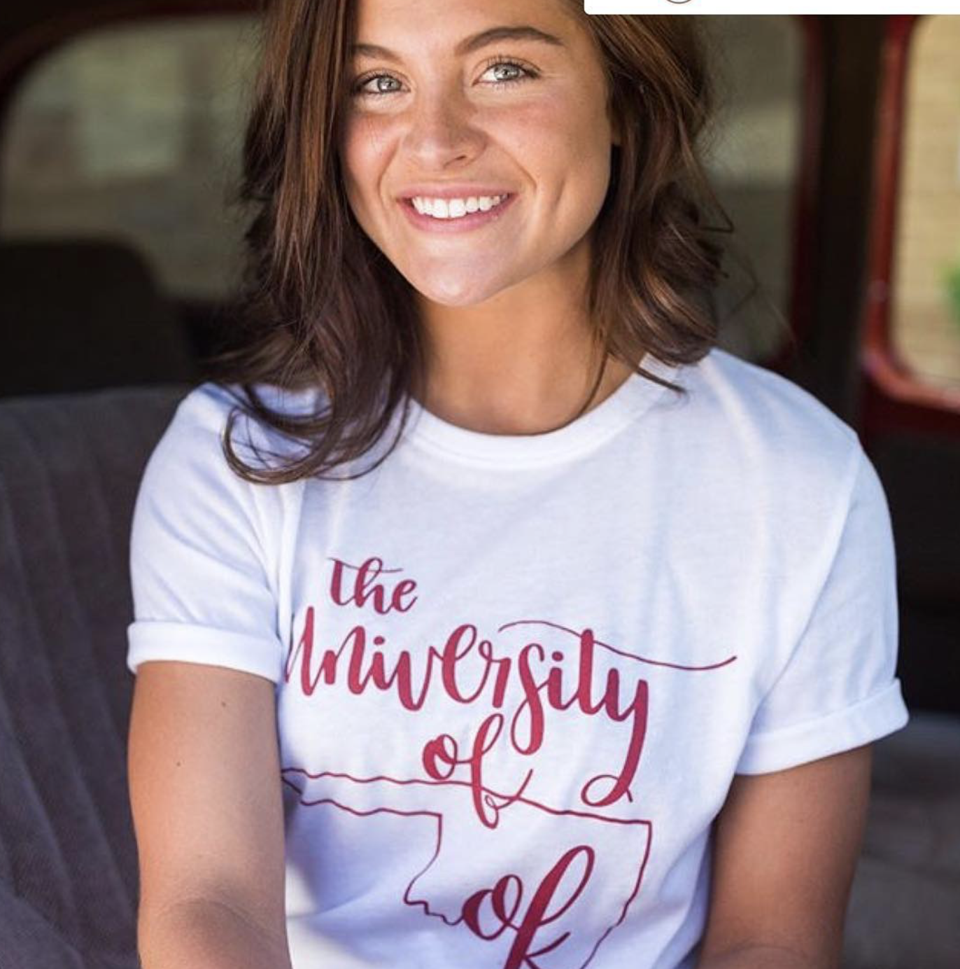 A woman with brown hair and blue eyes smiling, wearing a white t-shirt with red cursive text that reads 'the University of' and an outline of the state of Michigan.