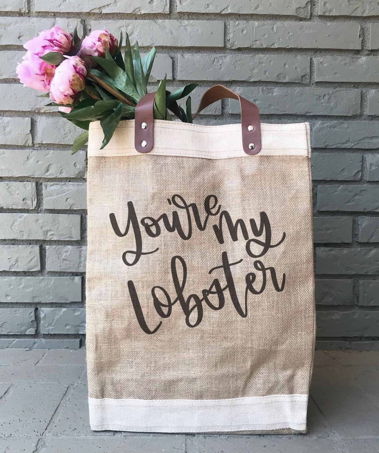 A tote bag with the words "You're my lobstar" written on it, filled with pink peony flowers, resting against a gray brick wall.