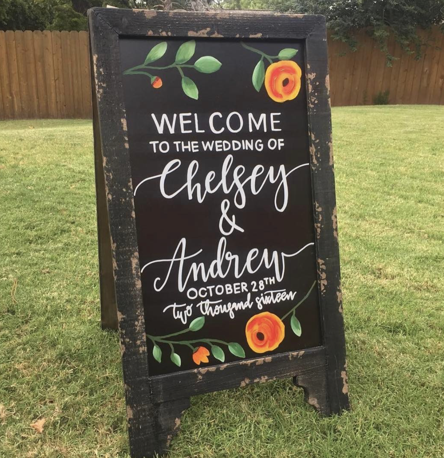 A black chalkboard wedding sign with floral decorations at the top and bottom, welcoming guests to Chelsea and Andrew's wedding on October 28th, two thousand sixteen.