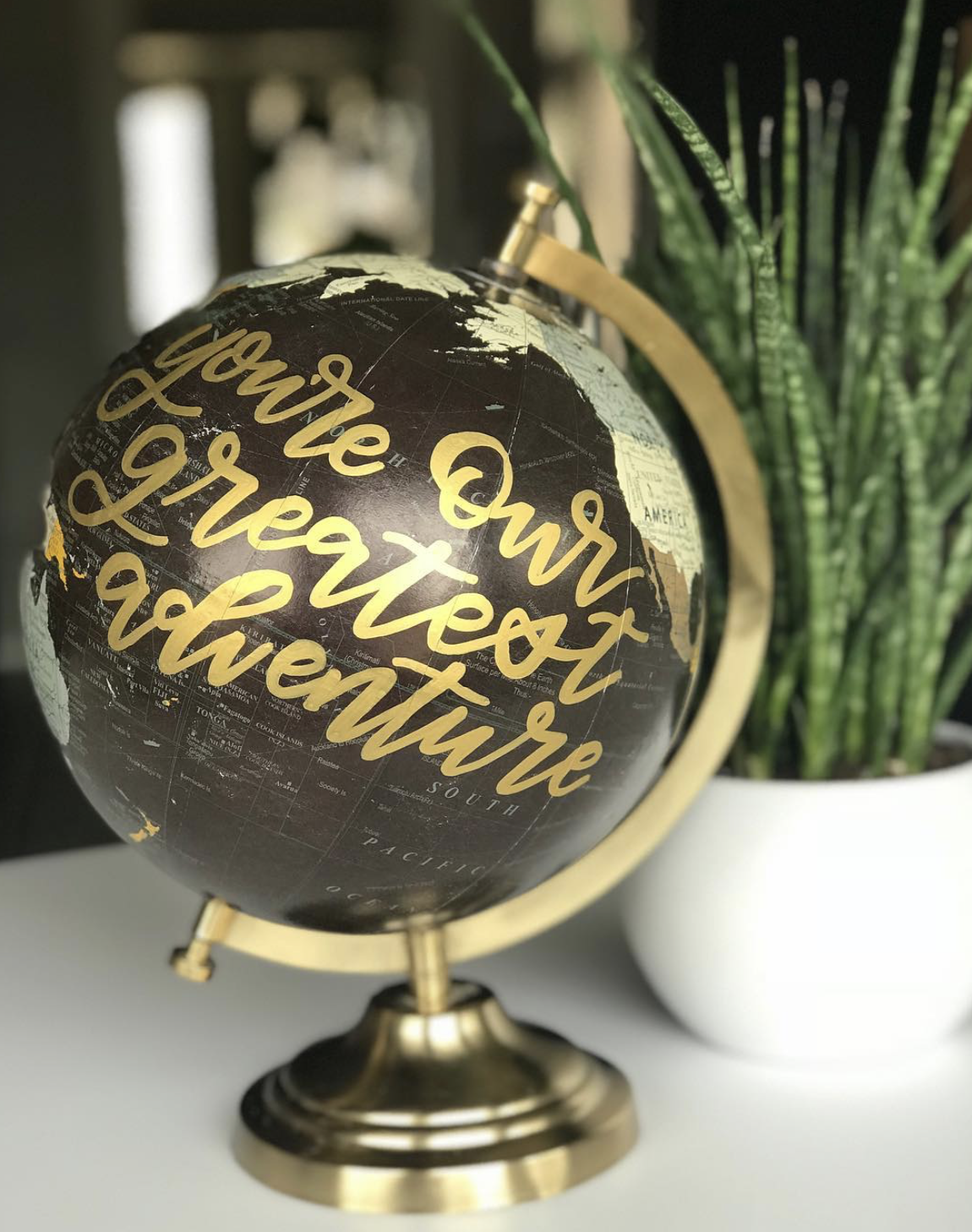 A black globe with gold text that reads 'Our Earth deserves attention', on a stand with a gold rim, placed on a white surface next to a potted green plant.
