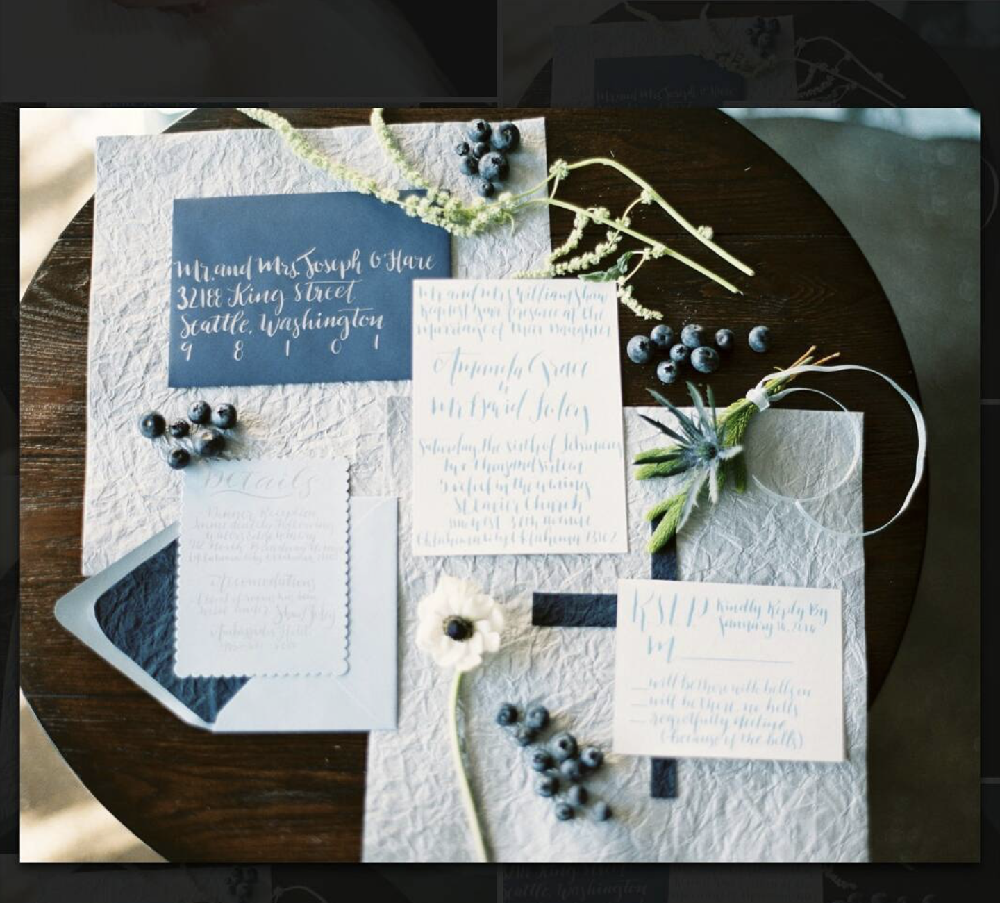 Flat lay of wedding invitation suite on textured white paper with navy, white, and floral accents, including invitation cards, RSVP card, envelope, white flower, and scattered blueberries on dark wooden table.