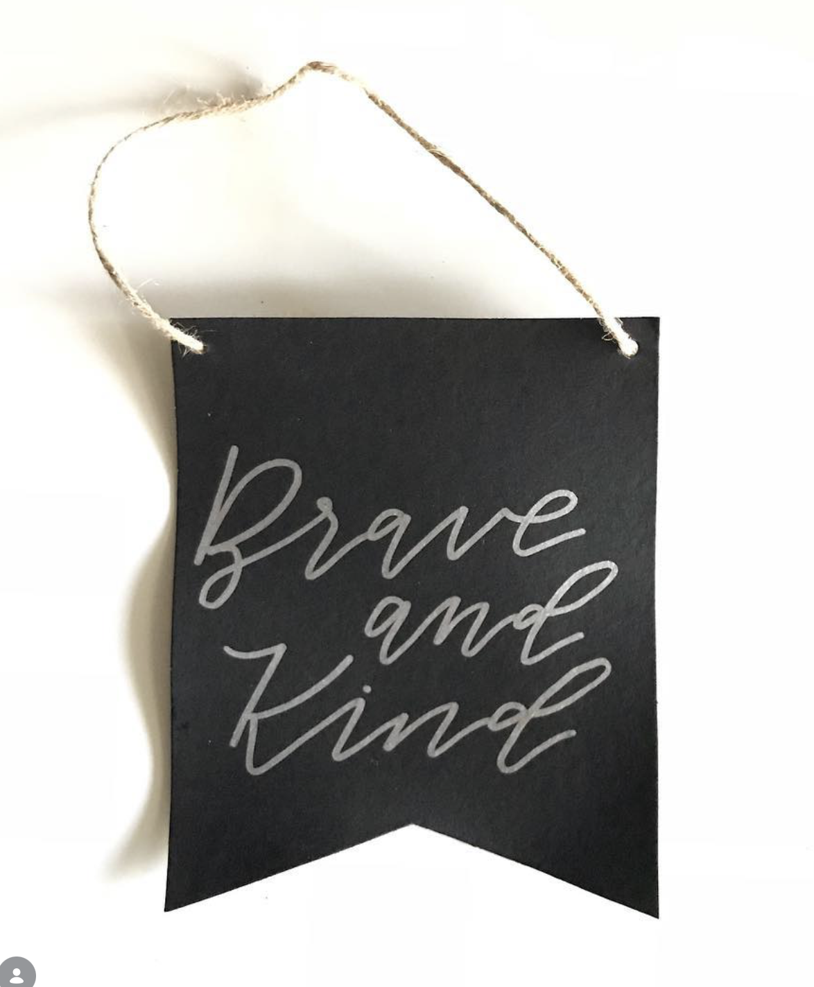 Black hanging sign with cursive writing that says 'Brave and kind,' attached with a string.