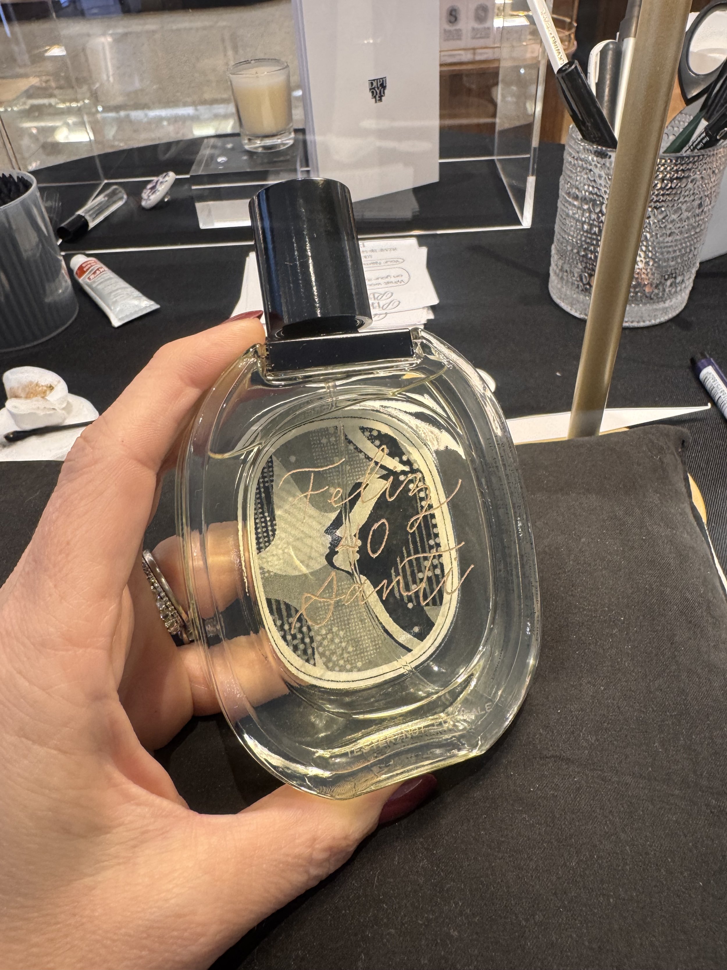 A hand holding a transparent plastic cup with clear liquid, with the word 'Fenty' on it, in a setting resembling a store or café with various items on a black table in the background.
