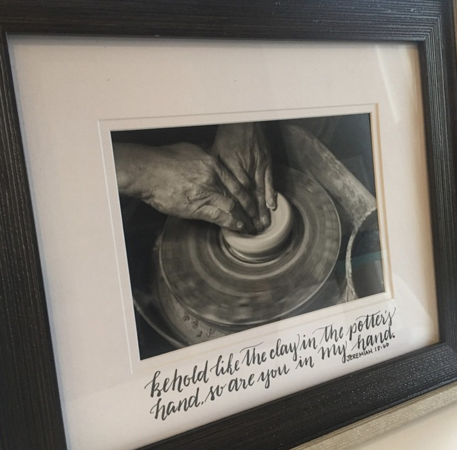 Framed black and white photograph of a hand shaping pottery on a pottery wheel with a quote at the bottom that reads: "I held like the clay in the potter's hand, so are you in my hand." - Jeremiah 18:6.