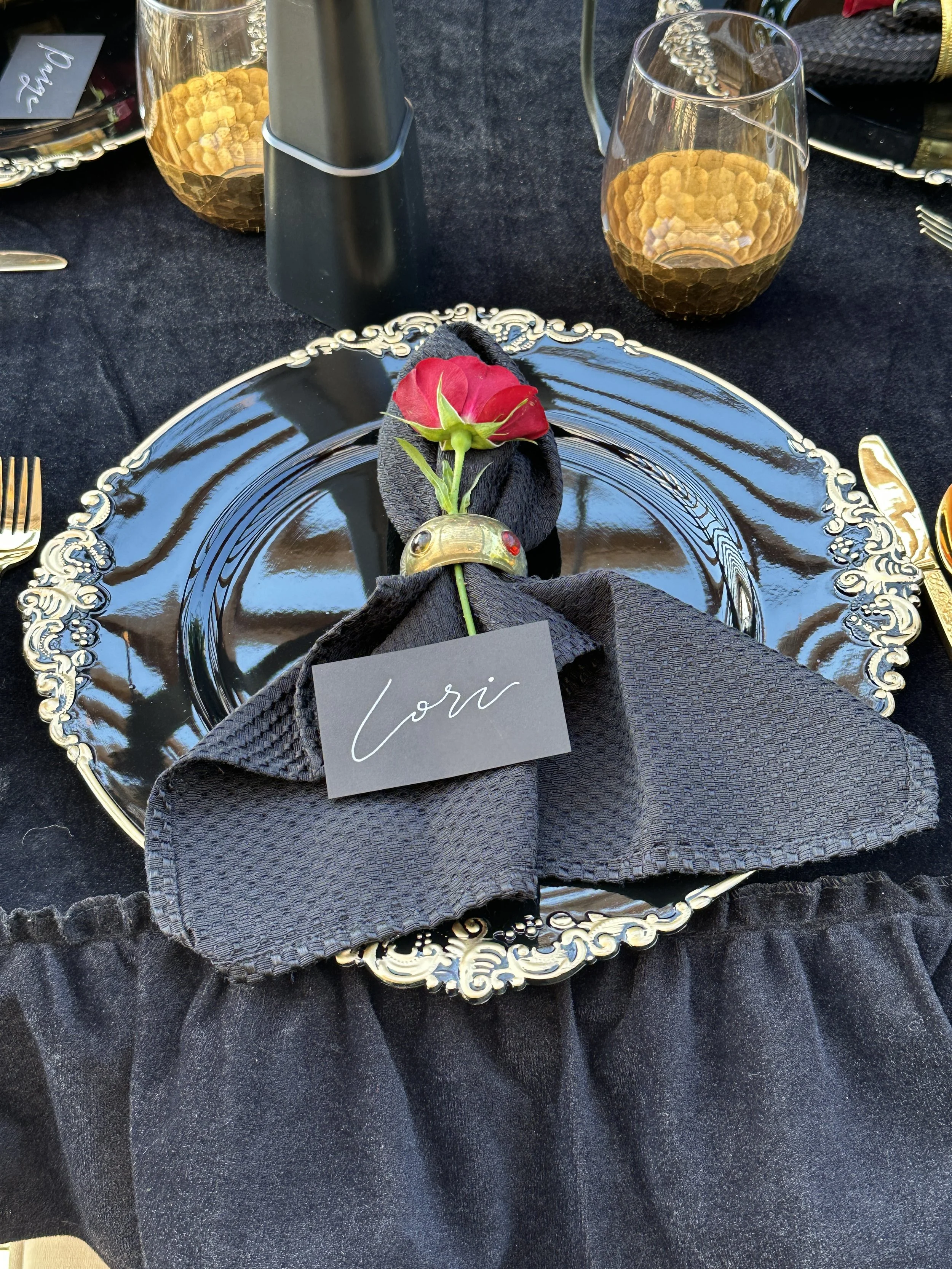 A table setting with a black ornate plate, black cloth napkin with a gold napkin ring, and a single red rose. There are two glasses with honey-colored drinks, a black bottle, and a silver tray with sweets in the background. The place card reads 'Lori'.