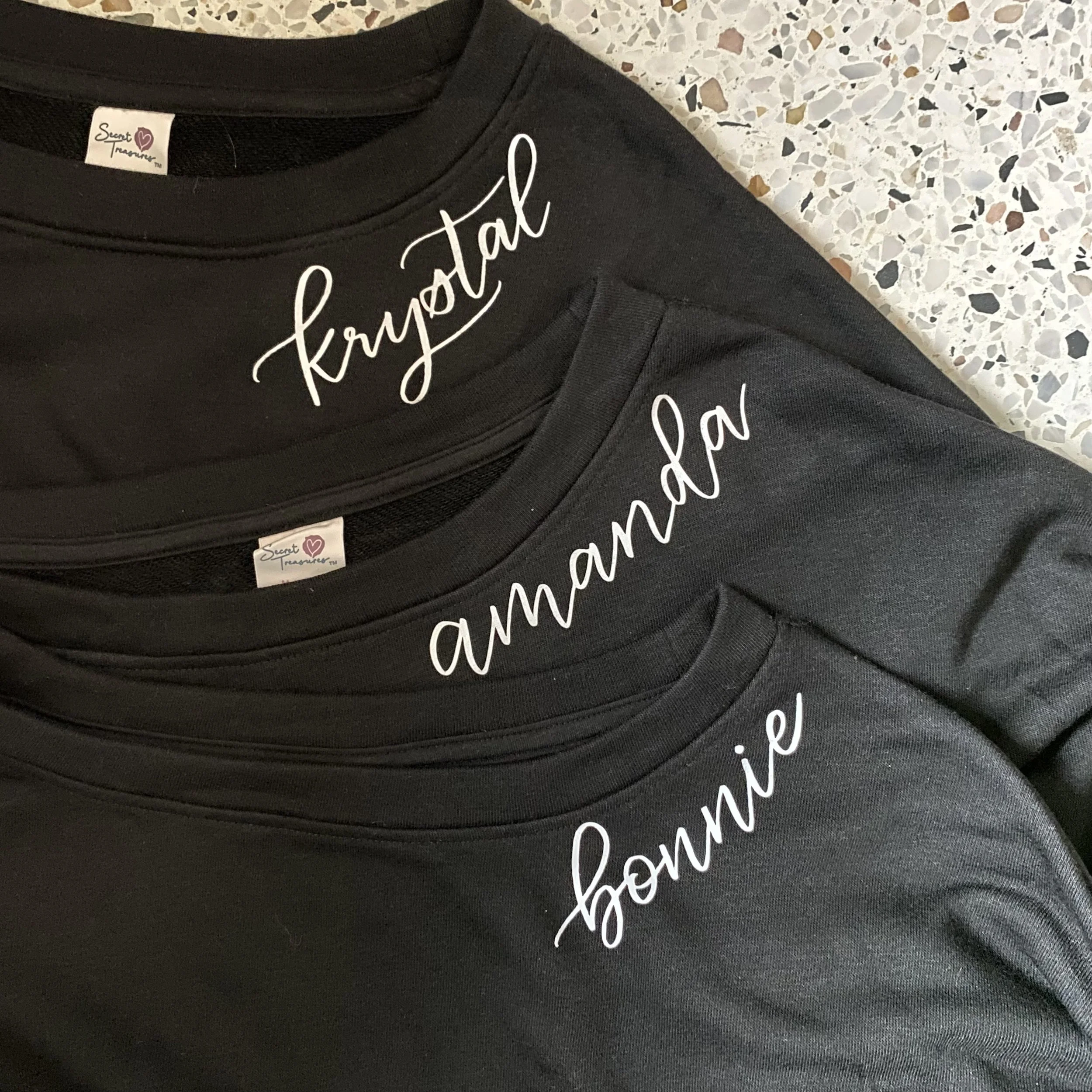 Close-up of a black T-shirt with white cursive writing and a small clothing tag that says 'Secret Treasures' with a heart symbol. The shirt is placed on a speckled terrazzo surface.
