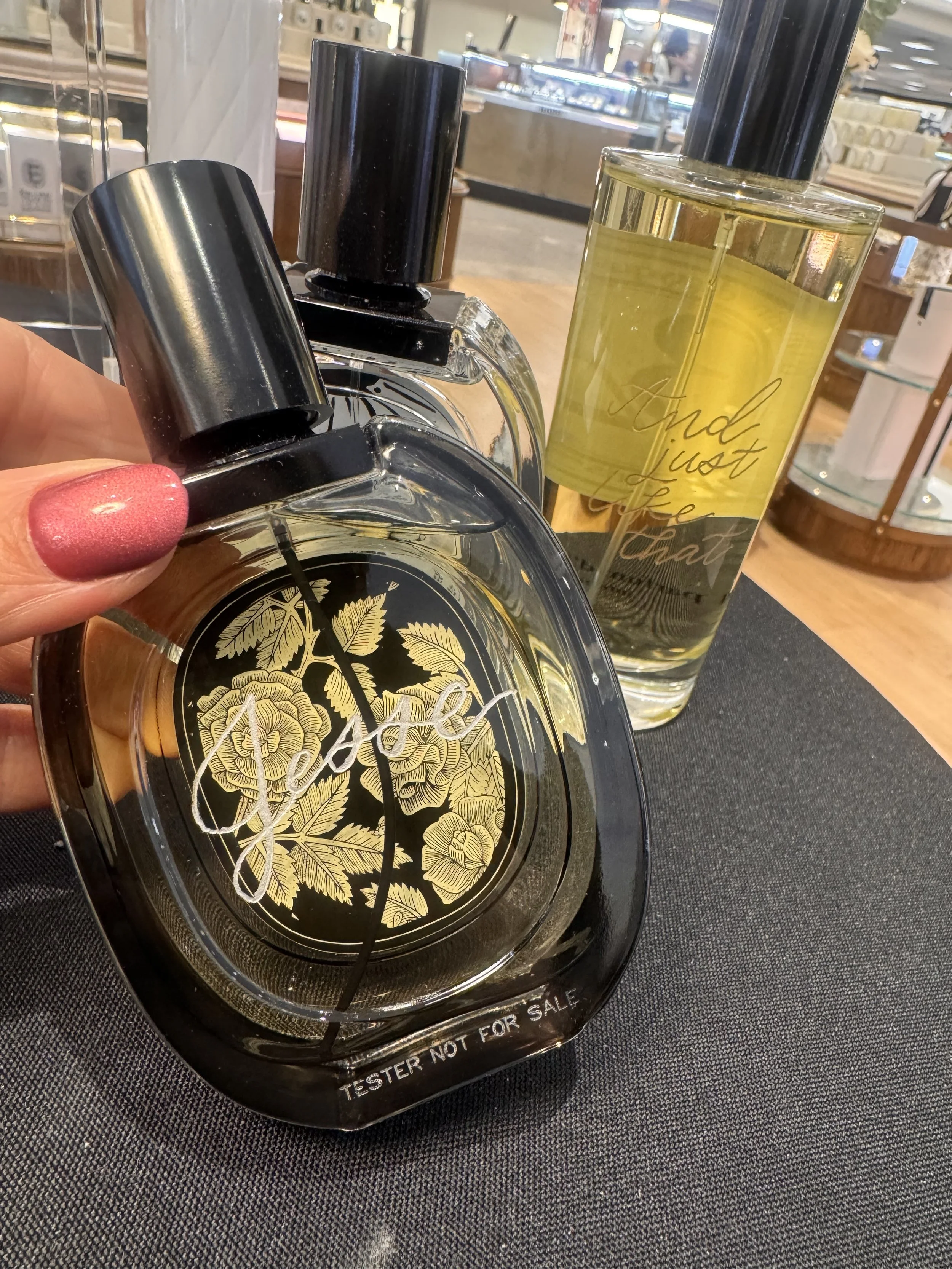 Close-up of a perfume bottle with black and gold design, a second perfume bottle with yellow liquid and black cap, and a person's hand with painted nails, set on a black surface in a store.