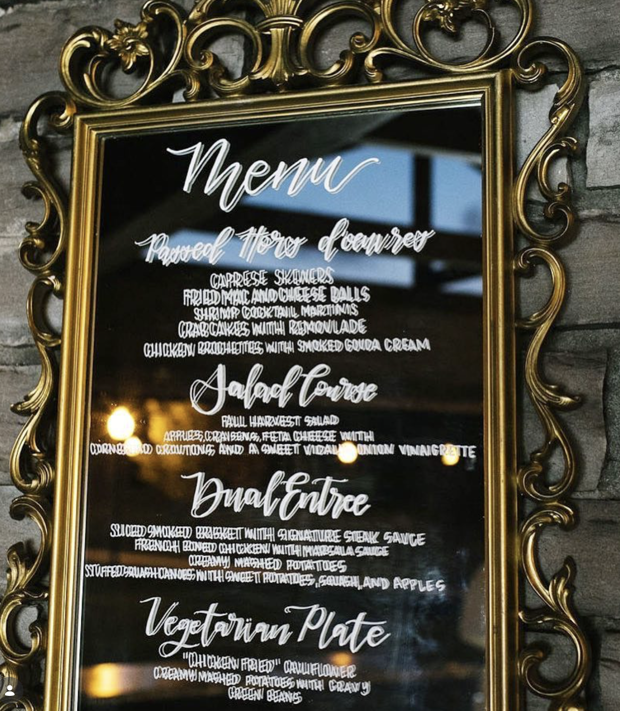 A decorative black menu board with gold ornate frame listing food items in white cursive font, taped to a stone wall. It shows categories for 'Passed Hors d'oeuvres,' 'Salad Course,' and 'Vegetarian Plate' with detailed descriptions of each dish.