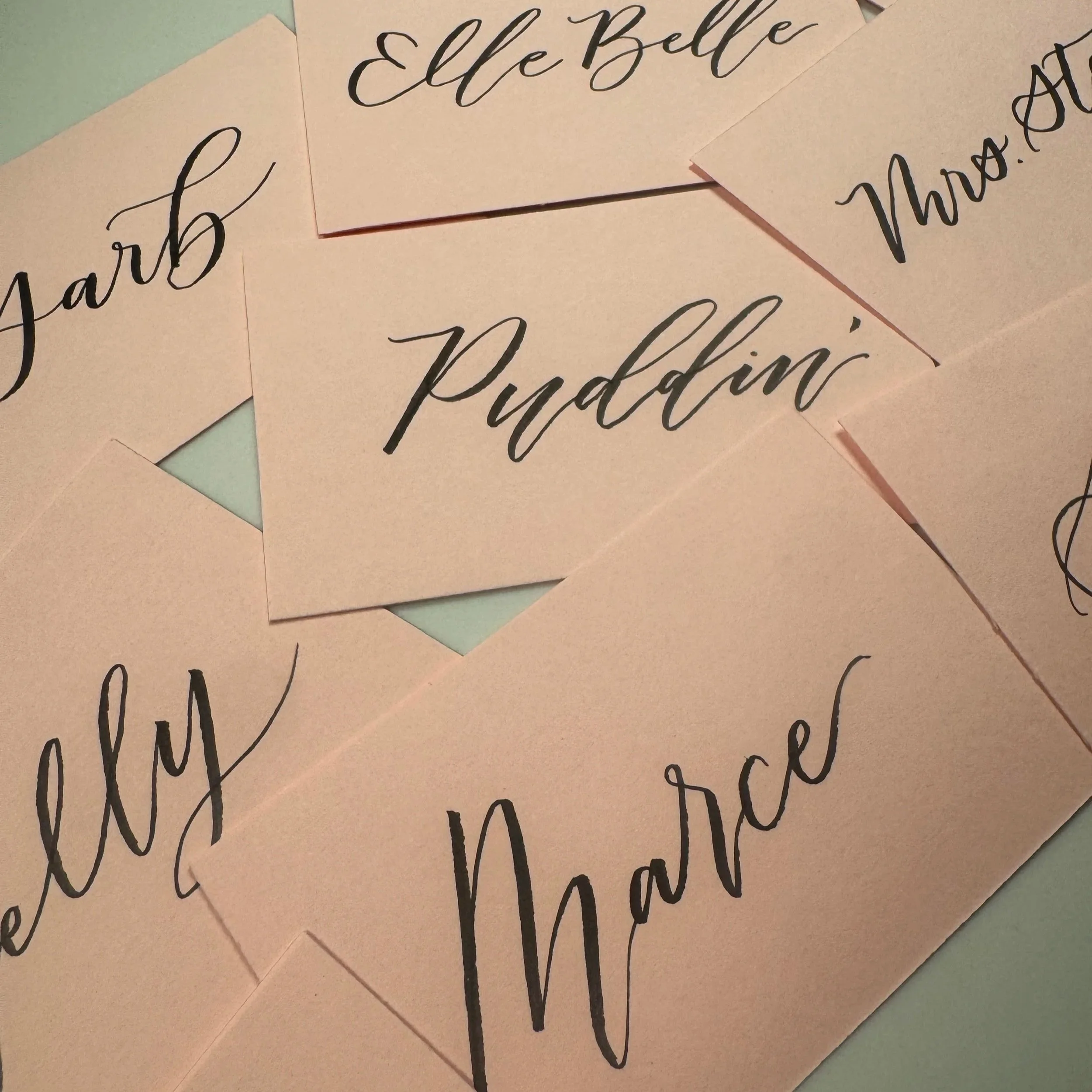 Close-up of several handwritten notes with cursive writing on pastel-colored paper, reading 'Elle Belle', 'Yard', 'Puddi...' and other incomplete words, with some notes overlapping.