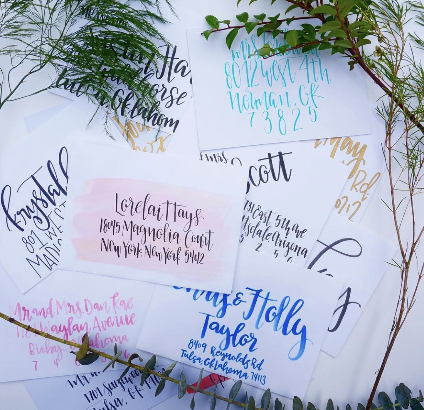 Several handwritten envelopes with addresses on them, decorated with sprigs of greenery, arranged on a flat surface for mailing or a wedding. The envelopes are in various colors and handwriting styles.