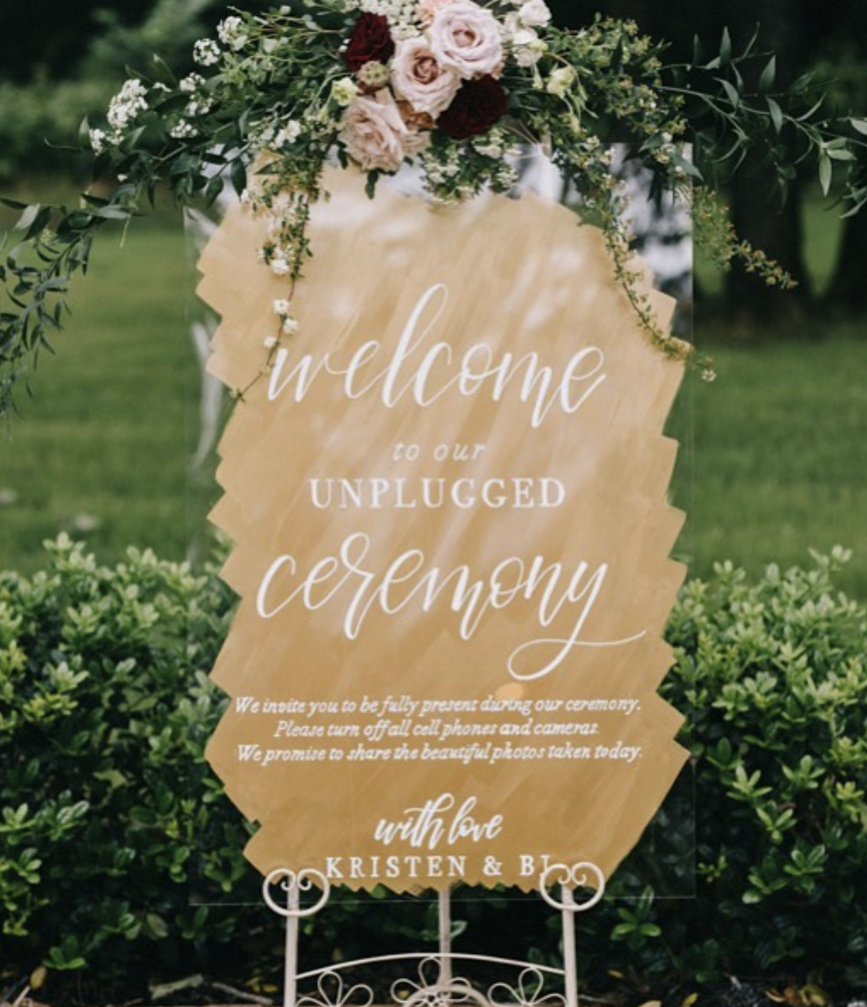 A wedding sign decorated with flowers and greenery, encouraging guests to be fully present at the unplugged ceremony, signed with love by Kristin and Bryan.