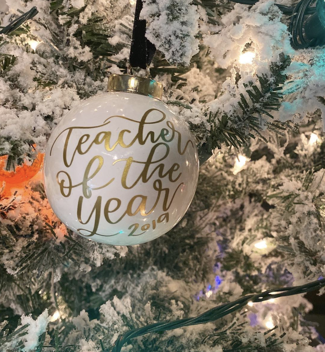 A white Christmas ornament with gold cursive text saying 'Teacher of the Year 2019', hanging on a snow-flocked Christmas tree with lights.
