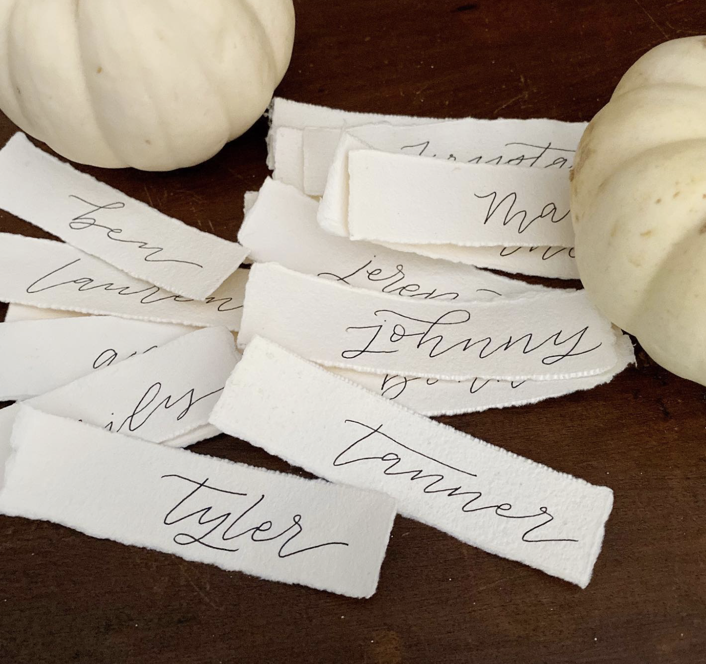 Several torn pieces of white paper with handwritten names such as Johnny, Taylor, and others, scattered on a wooden surface, with two white pumpkins in the background.
