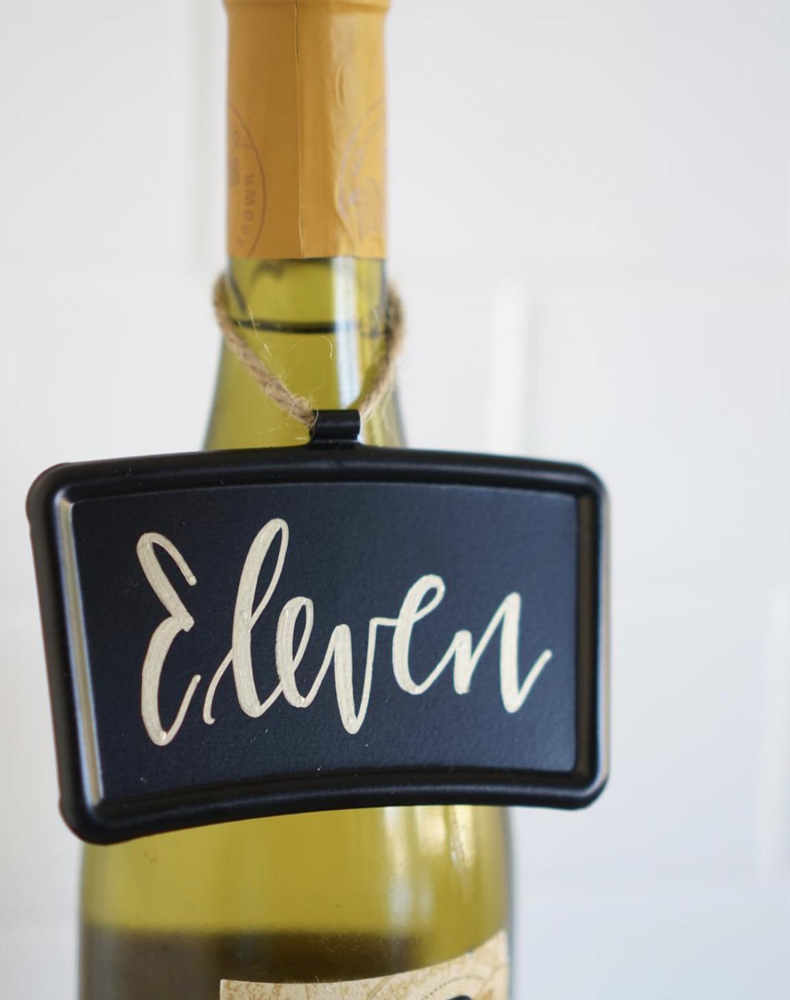 A bottle of champagne with a black chalkboard label that reads 'Eclewm', hanging on the neck with a piece of string.