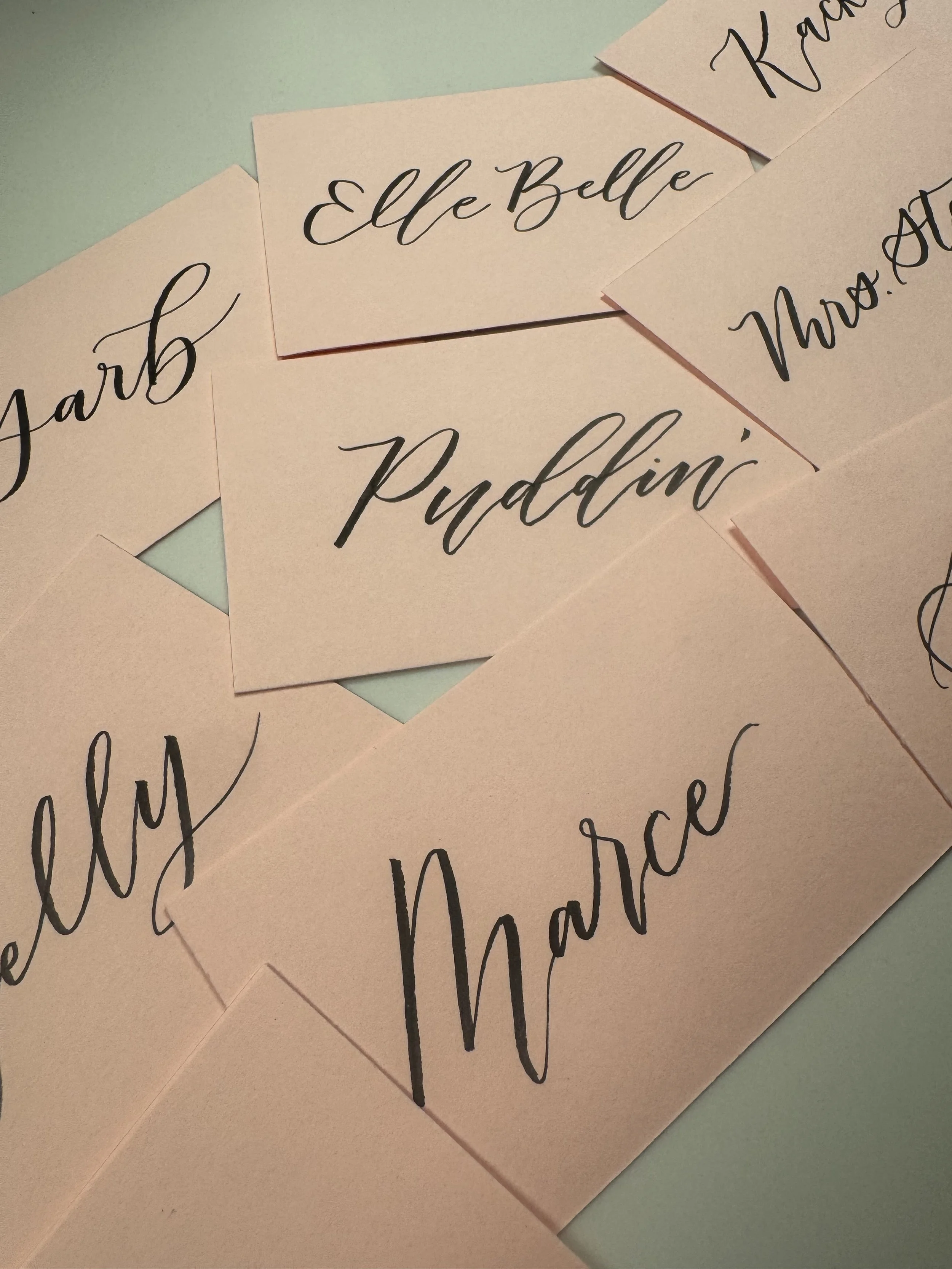Several pink cards with elegant black handwritten calligraphy text, with words like 'Elle Belle,' 'March,' 'Paddini,' and 'Thanks' visible.