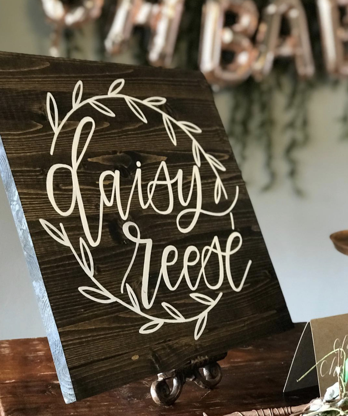 A wooden sign with white cursive text that reads 'day ~ of' surrounded by a decorative leafy wreath.