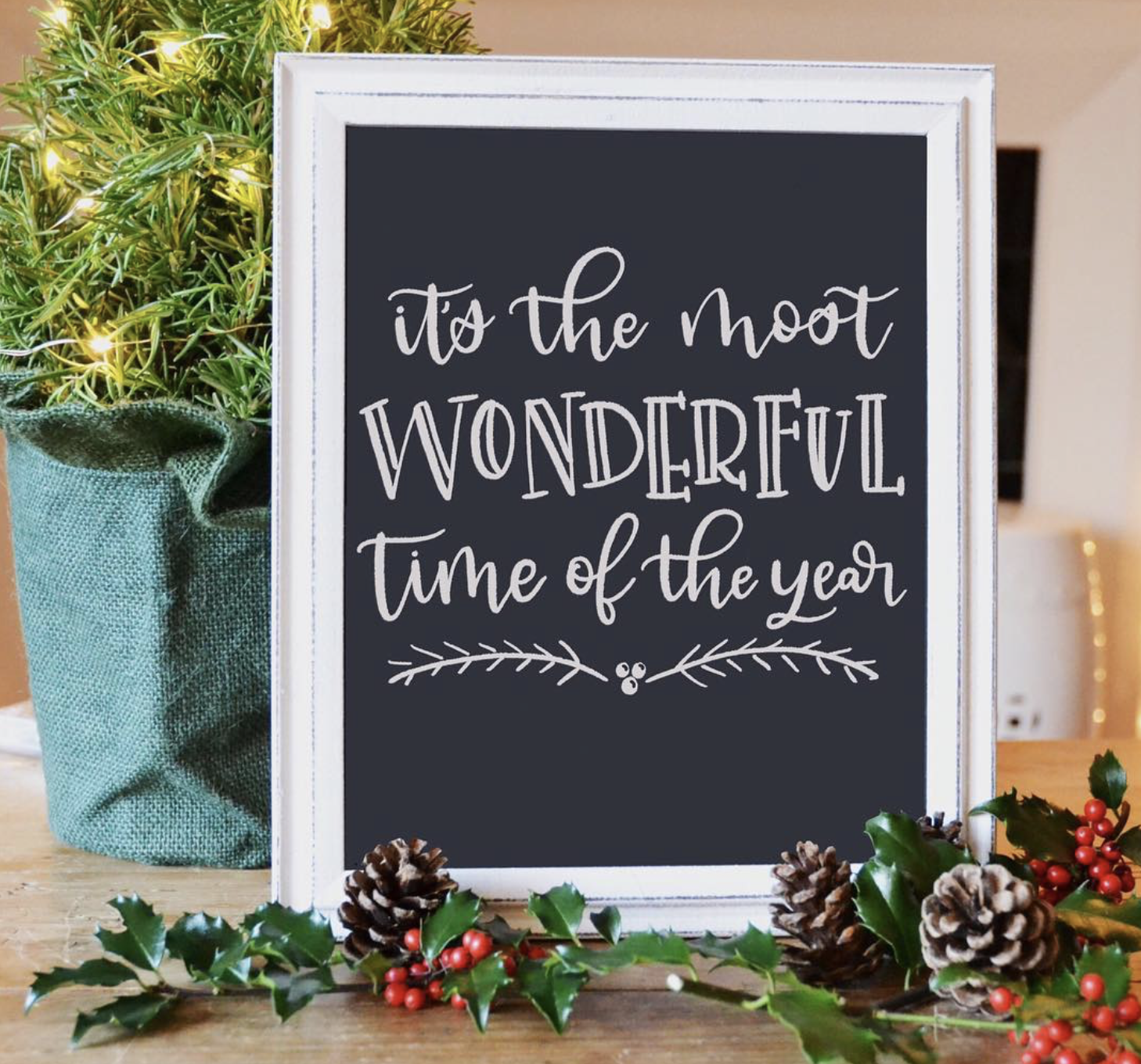 A framed festive sign with the words "it's the most WONDERFUL Time of the year" written in white script on a dark background. The sign is decorated with holly and surrounded by pinecones and holly berries, with a Christmas tree decorated with lights 