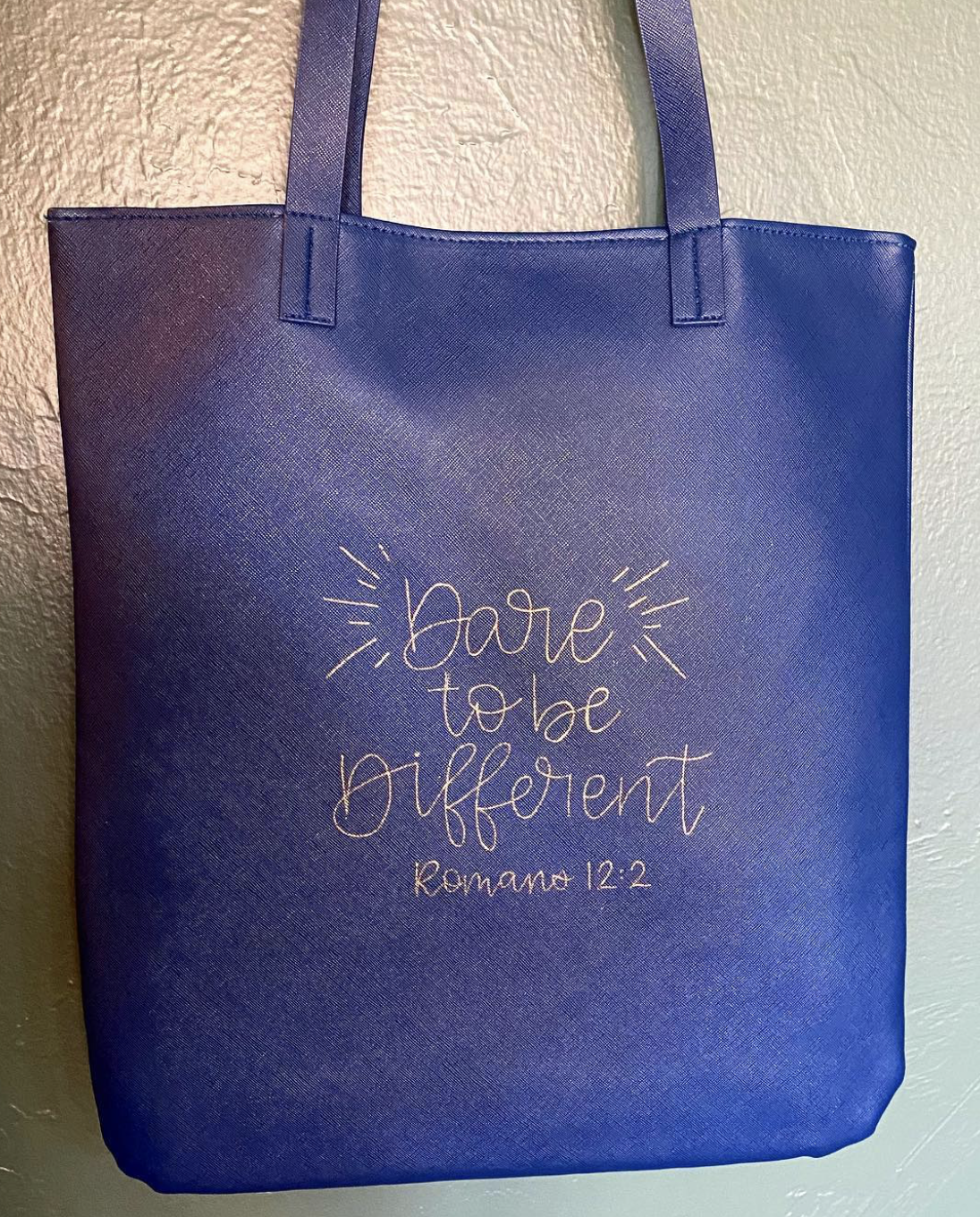 A blue tote bag with the words "dare to be different" and a reference to Romans 12:2 written on it.
