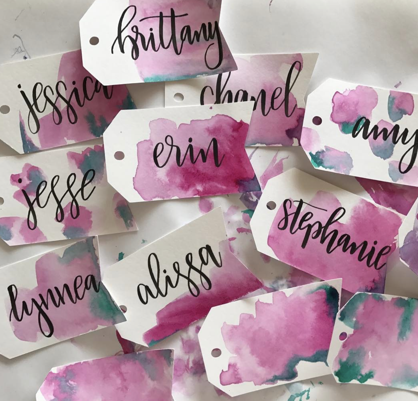 Multiple name tags with artistic pink, purple, and blue watercolor backgrounds, each handwritten with black ink in cursive. Names visible include Brittany, Jessica, Erin, Amy, Stephanie, Alyssa, and Lianne.