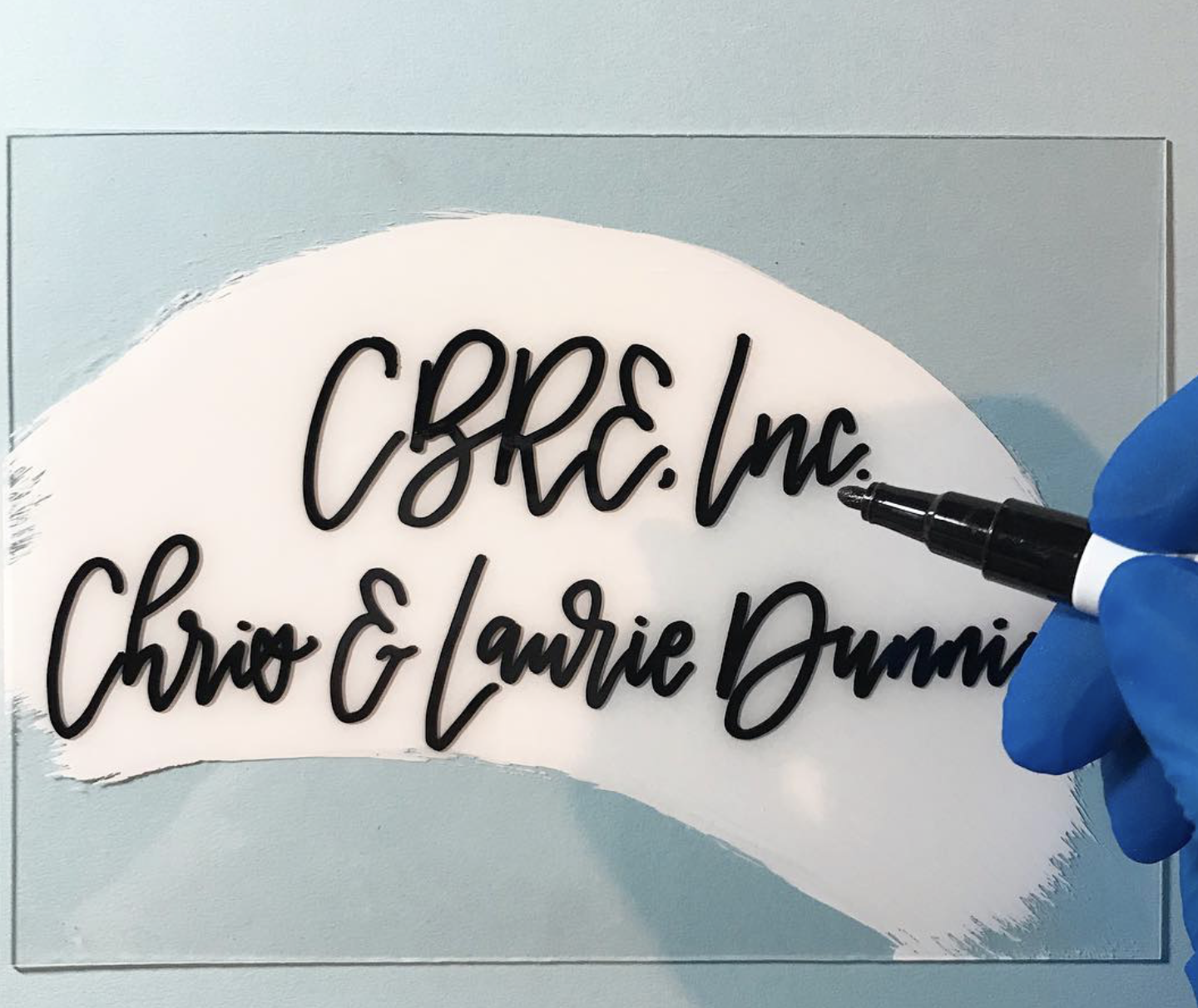 Close-up of a person in blue gloves writing on a sign with a black marker, the sign has the text 'C.B.C. Inc. Chiro & Lurie Dummi' in black cursive font on a white background with a decorative edge.