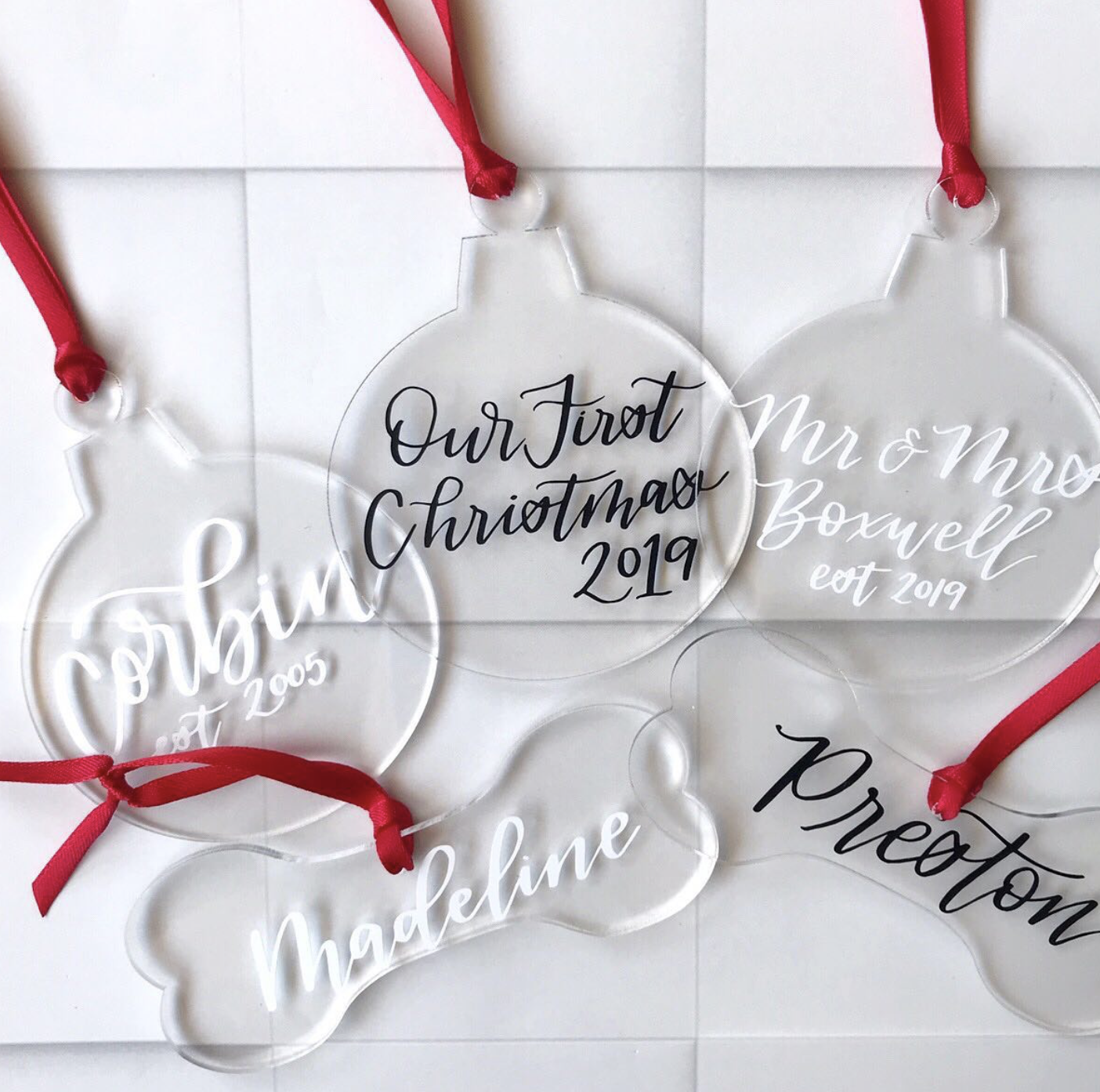 Clear holiday ornaments with black and white and black text and red ribbons, celebrating a family's first Christmas in 2019, with details of family members' names and years.