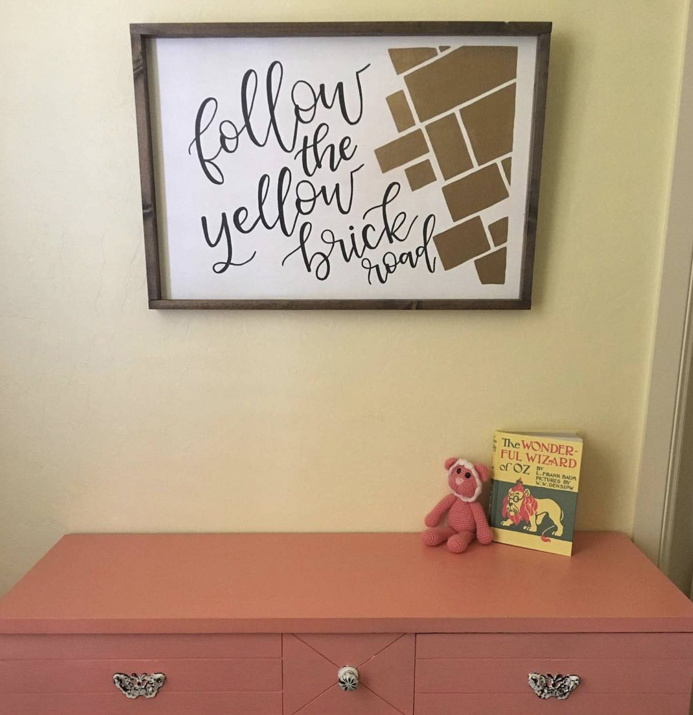 A wall decor sign with the phrase "follow the yellow brick road" above a pink dresser. On the dresser, there is a small pink knitted teddy bear, a book titled "The Wonderful Wizard of Oz," and a small white decorative knob. The wall color is light ye