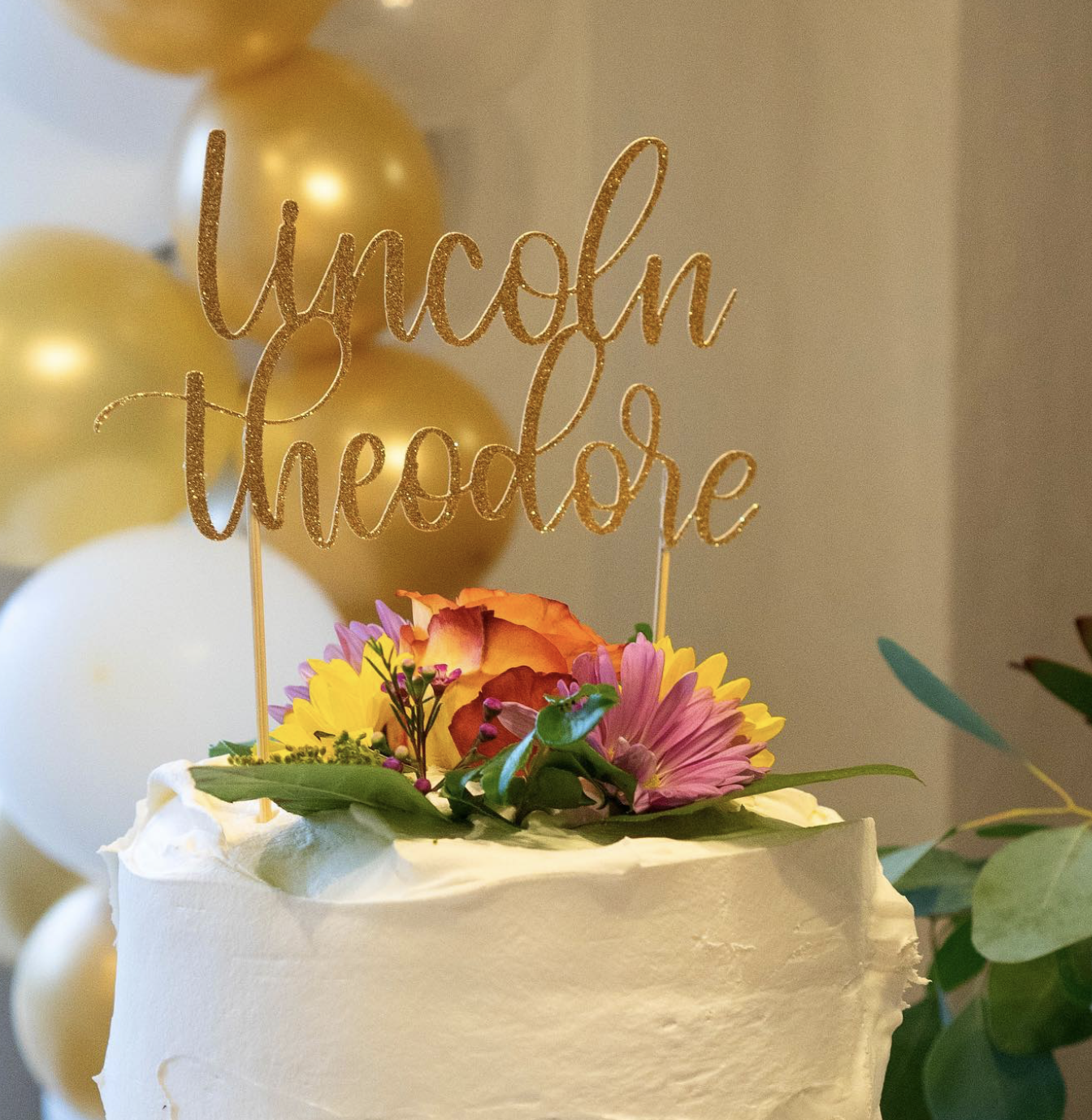 A white birthday cake decorated with fresh flowers and a gold glittery topper that reads "you got this". In the background are gold and white balloons.