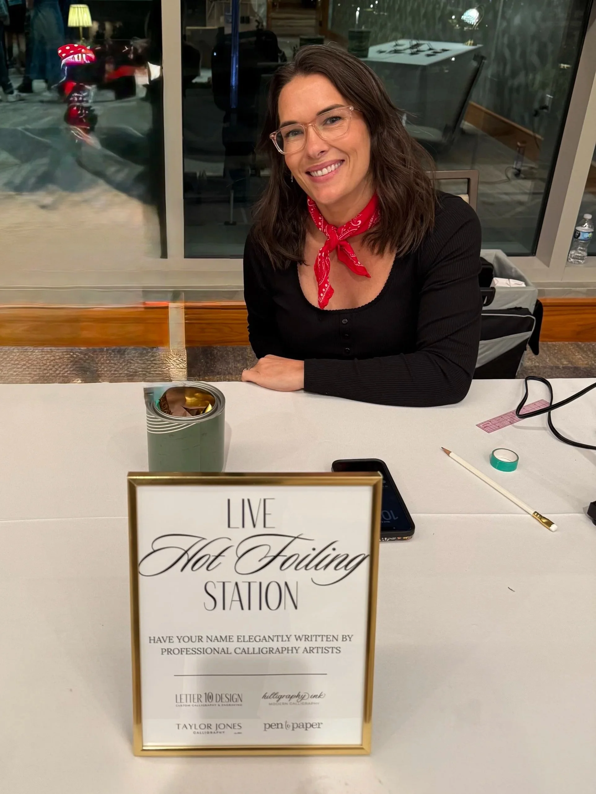 A woman sitting at a table with a sign that reads 'Live Hot Foiling Station'. The table has a pen, a roll of tape, a smartphone, and a container. Behind her, there is a large window showing an indoor space with people and objects, including a foiling machine.