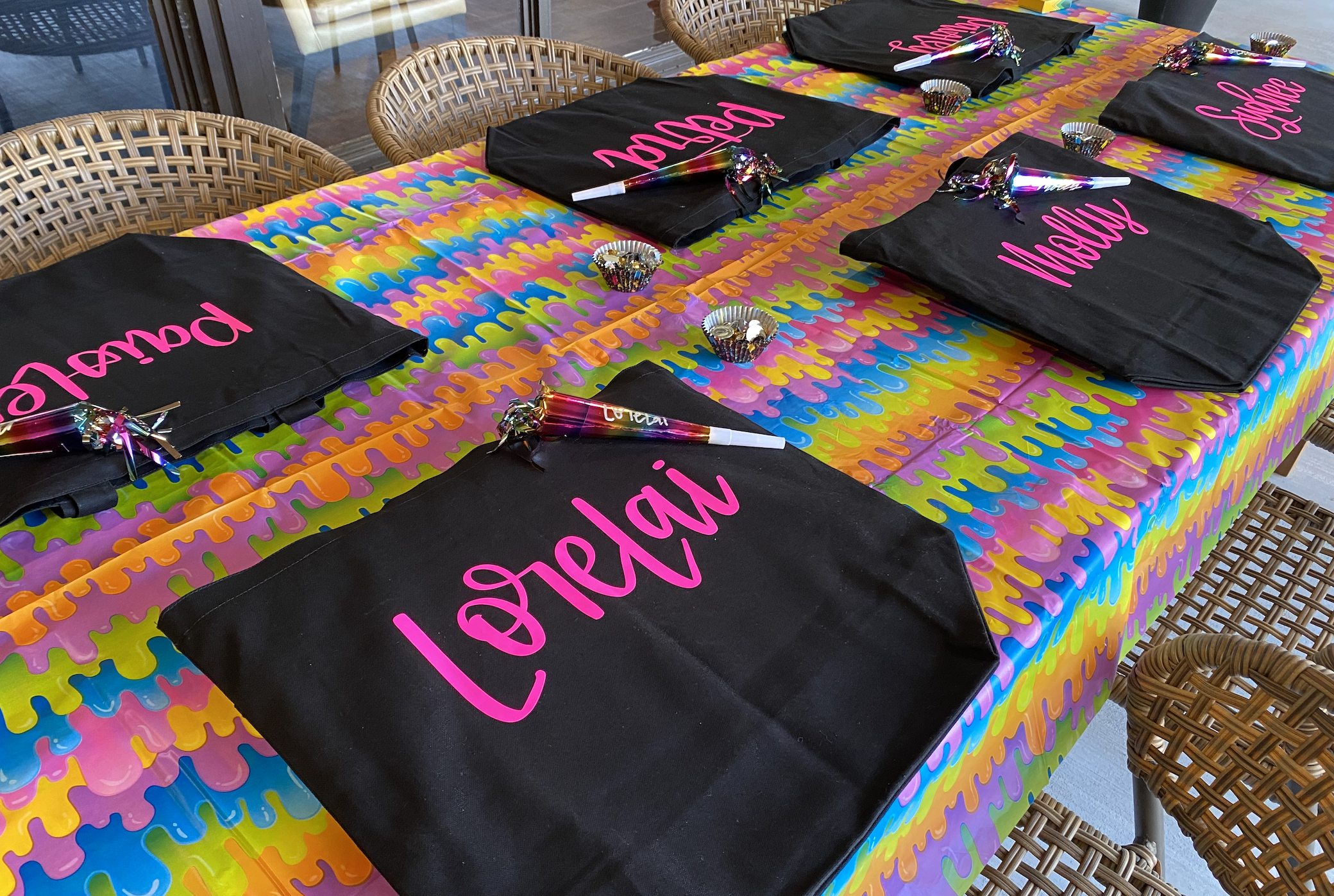 A party table decorated with a colorful, wavy-patterned tablecloth, four black gift bags with pink 'birthday' lettering, and party hats with shiny, multicolored fringe.