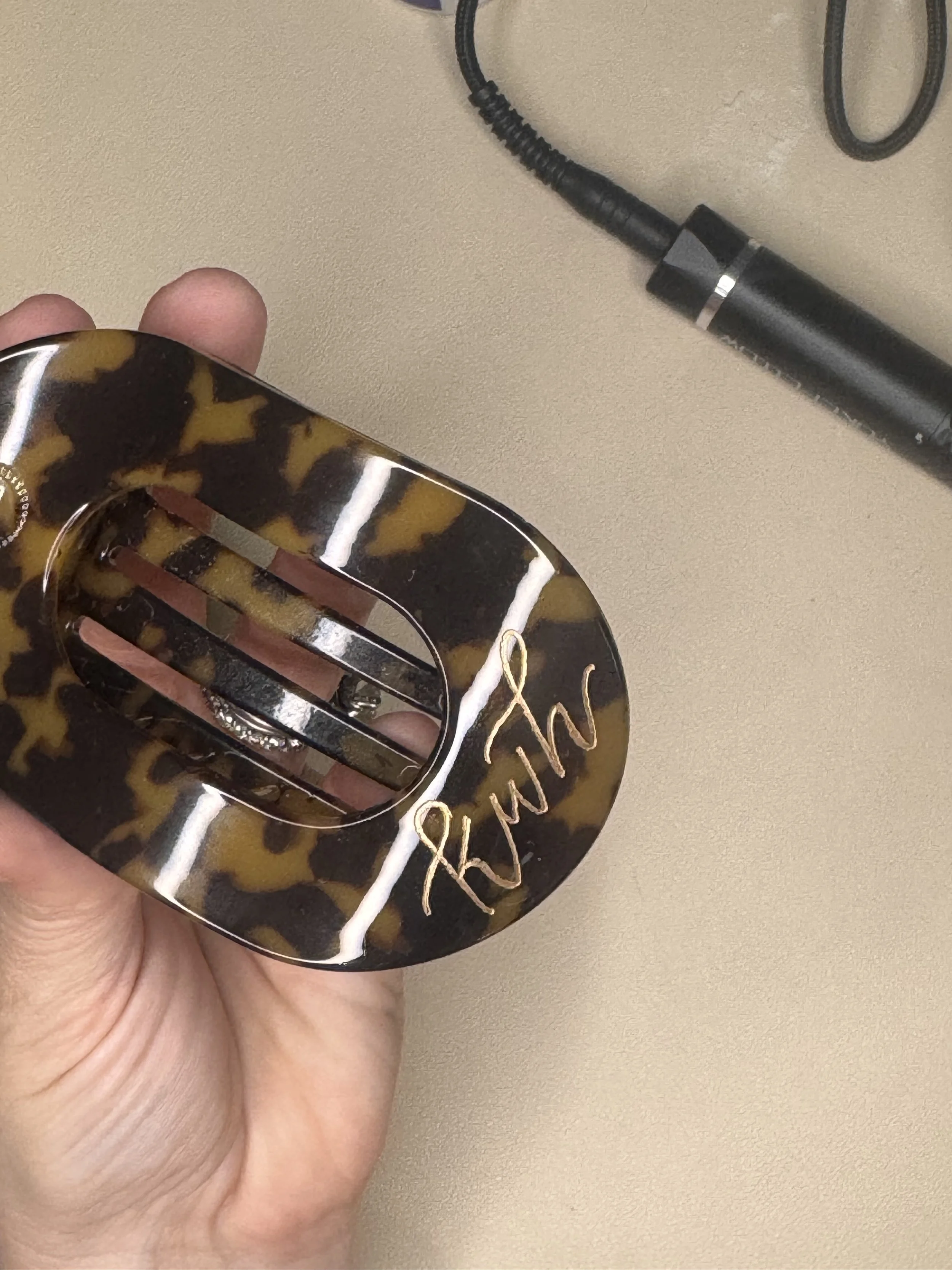 A person's hand holding a tortoise-shell style hair clip with gold accents, and a black portable microphone on a beige surface.